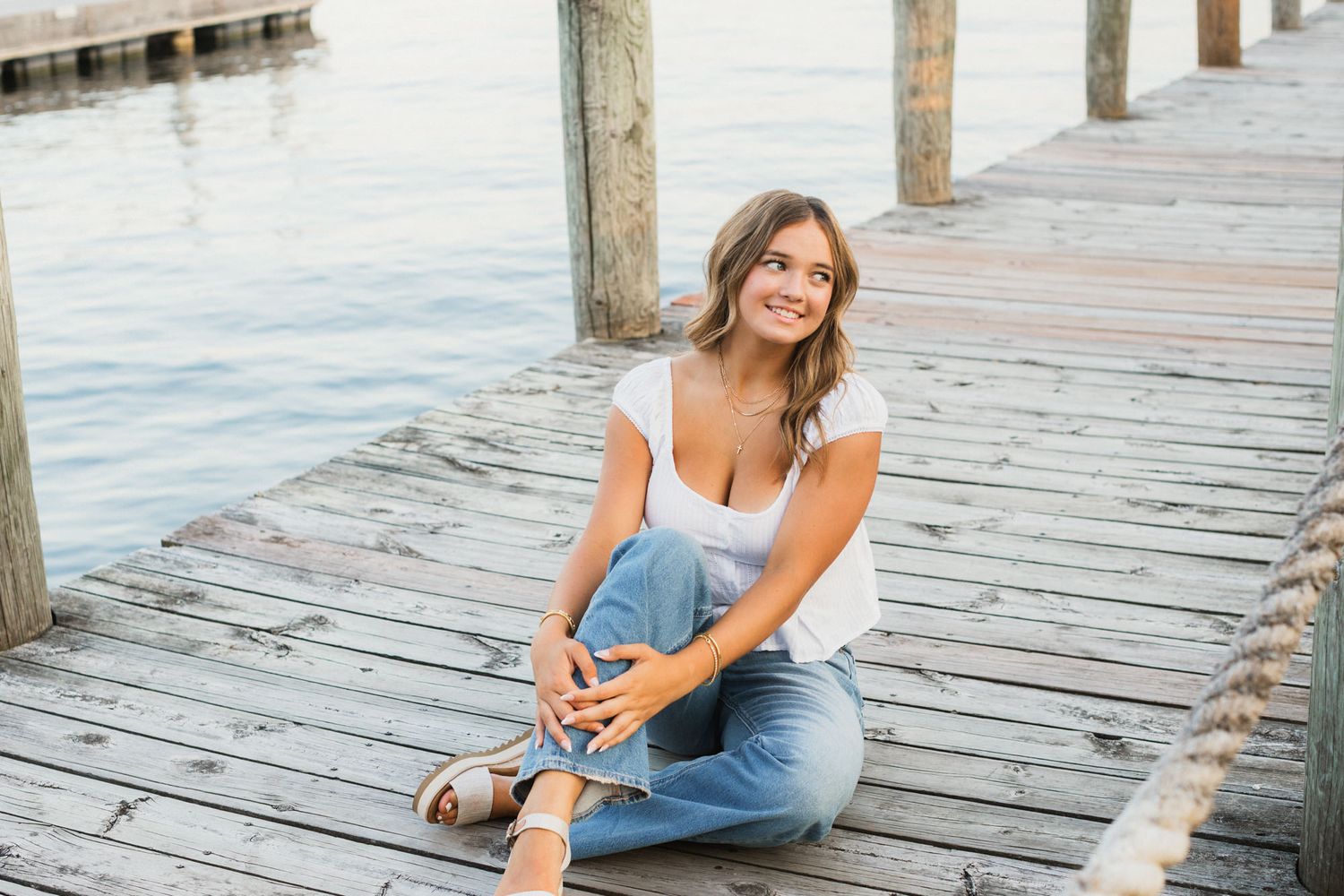 Young woman in white top and jeans sitting on a wooden dock by the water, smiling outdoors.