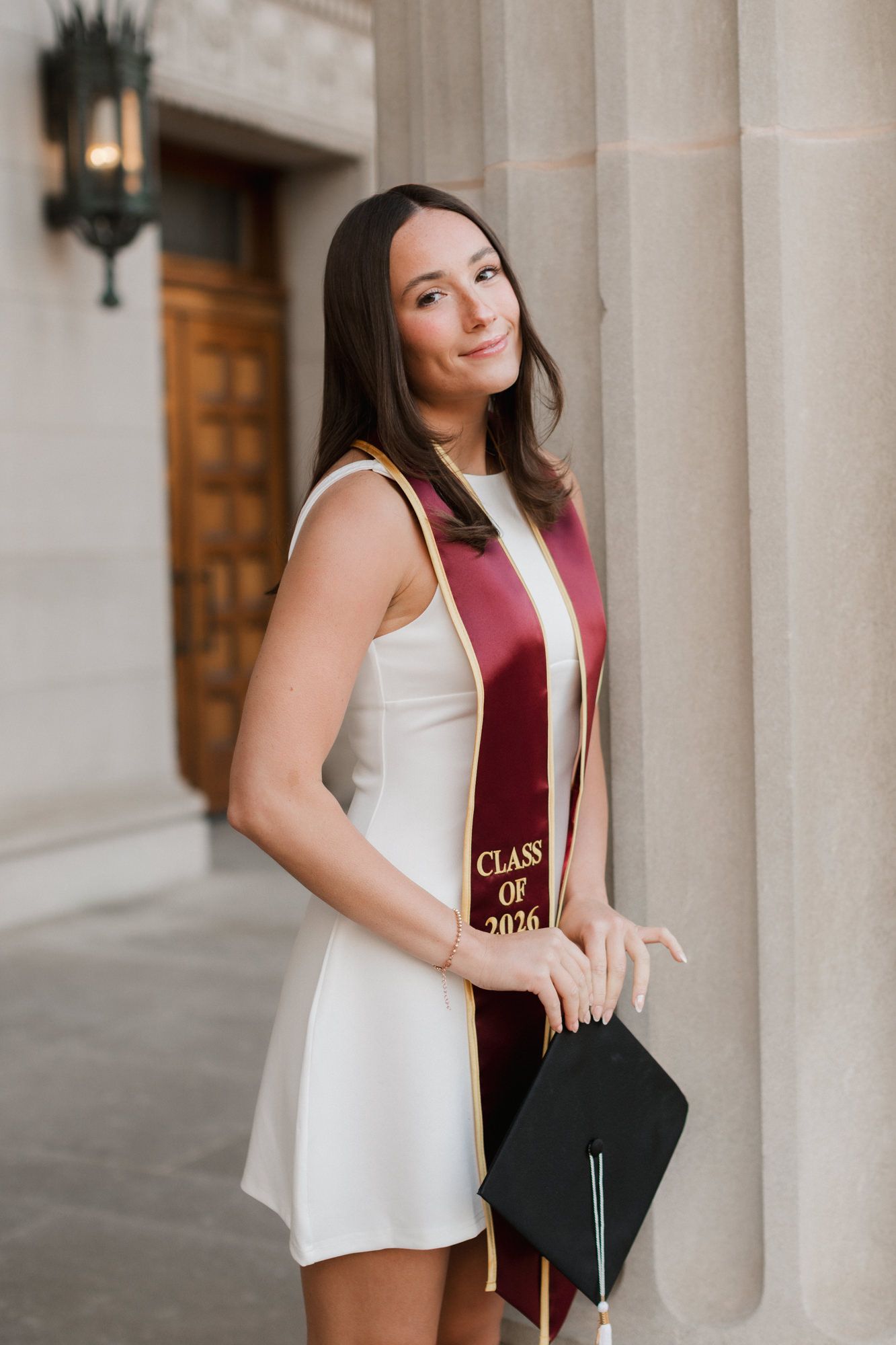 University of Minnesota Class of 2026 graduate in white dress and maroon stole posing with cap by stone columns.