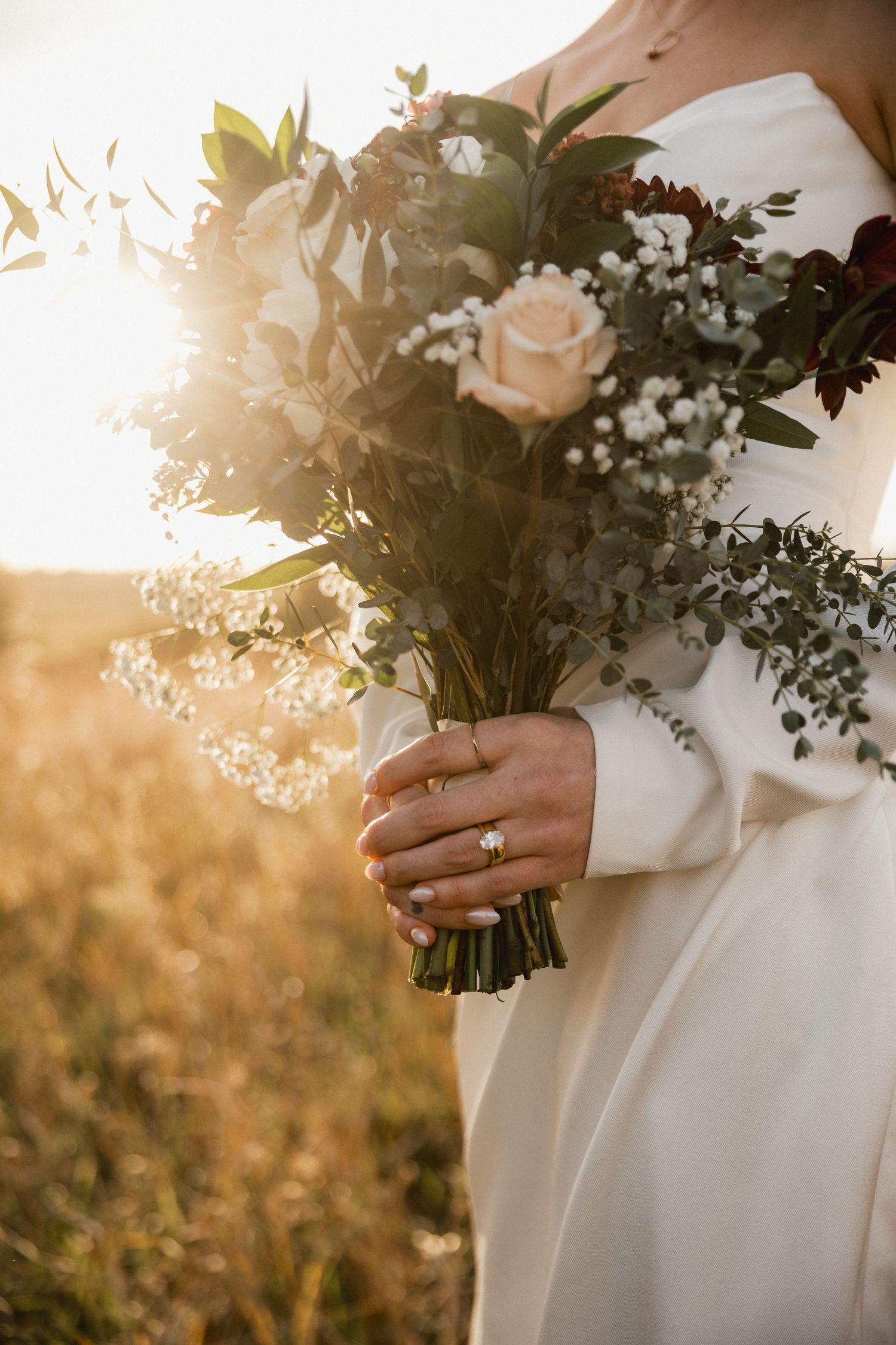Bride in a white gown holds a rustic wedding bouquet of cream roses, baby's breath and eucalyptus in warm golden sunlight.