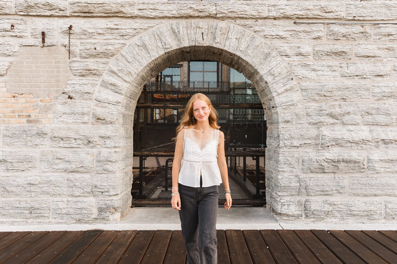 Fashion portrait under a stone archway on a wooden deck with textured limestone walls.