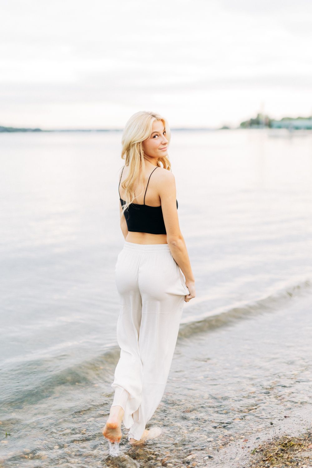 Woman in black top and flowing white skirt walks gracefully along a serene beachfront at sunset.