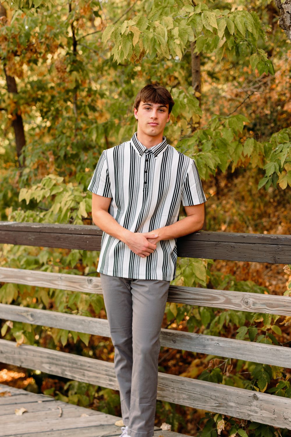 A person in a striped button-up shirt leans against a wooden fence railing in an autumn outdoor setting.