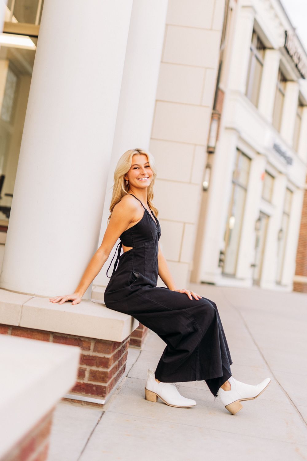 A person in a black dress and white sneakers poses casually on a brick ledge near white columns.