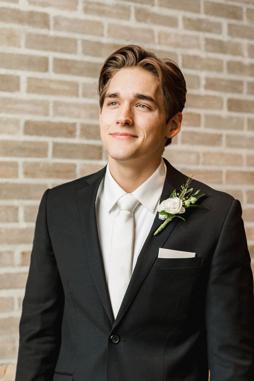 Wedding portrait of groom in classic black tuxedo with white boutonniere against brick wall.
