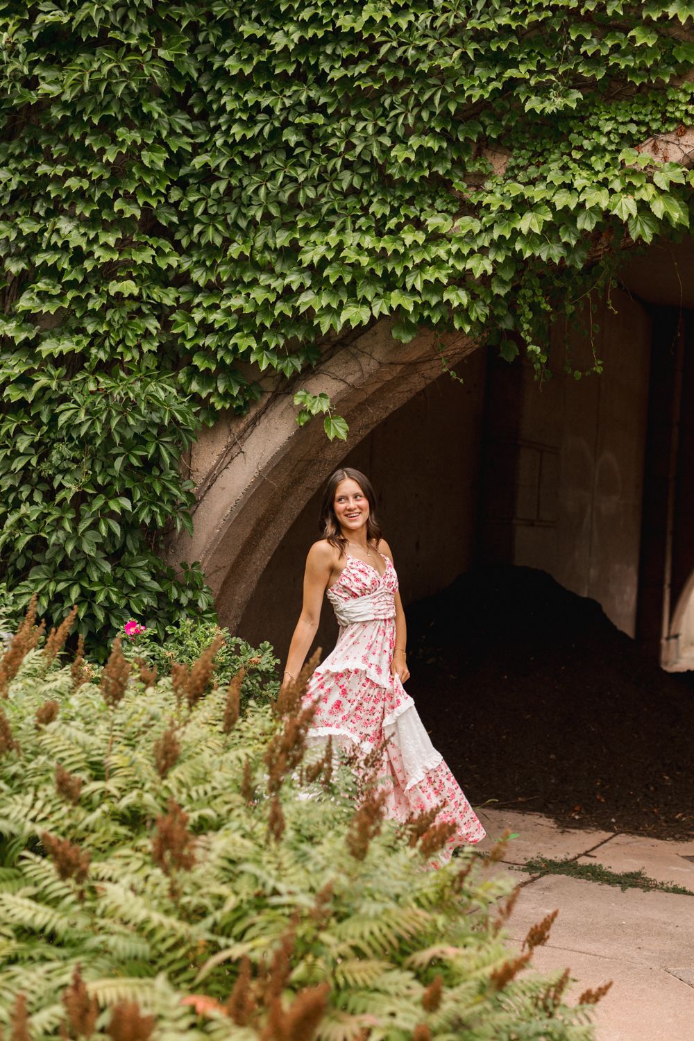 Someone wearing a floral sundress poses playfully among lush green ivy and ferns in an archway setting.