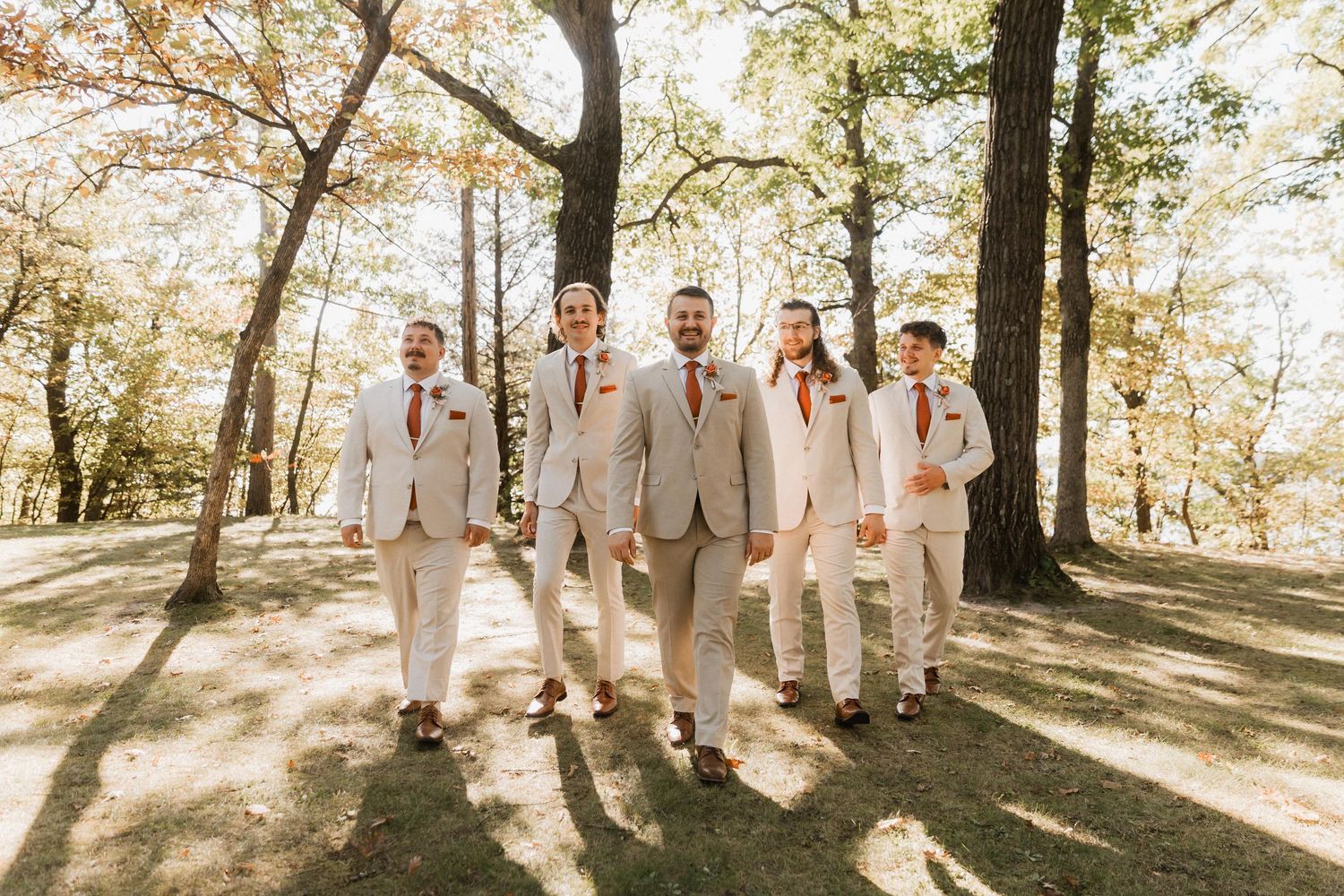 Wedding party in light beige attire poses together in a sun-drenched forest setting during golden hour.