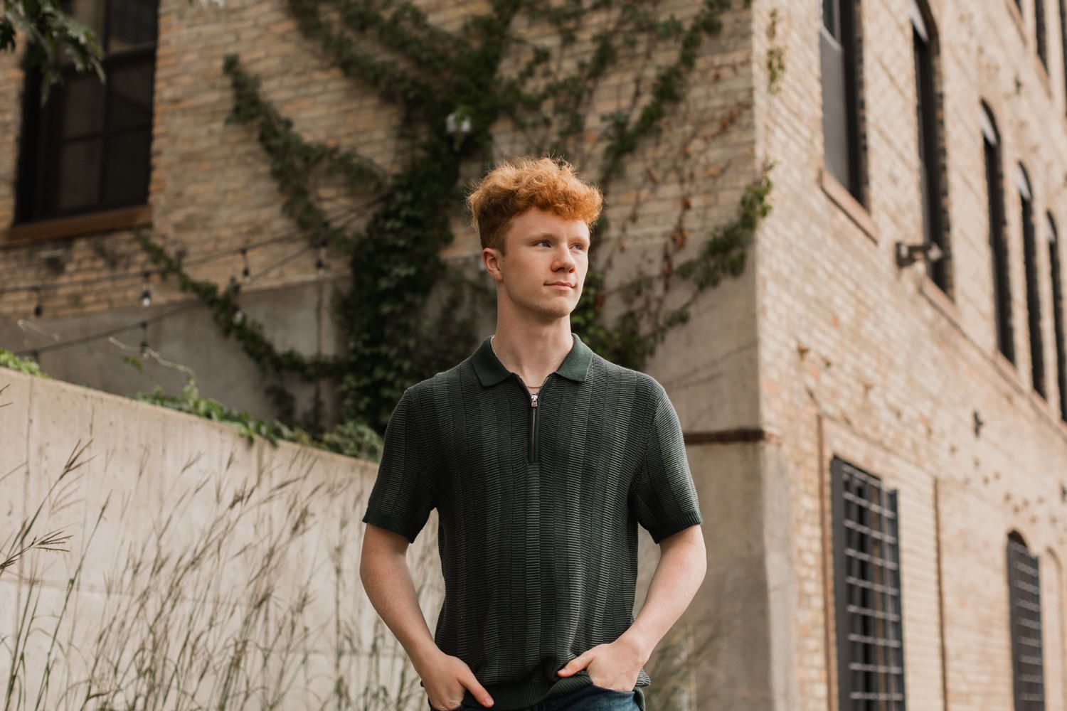 A person wearing a black shirt stands against a rustic brick wall with overgrown vines and foliage.
