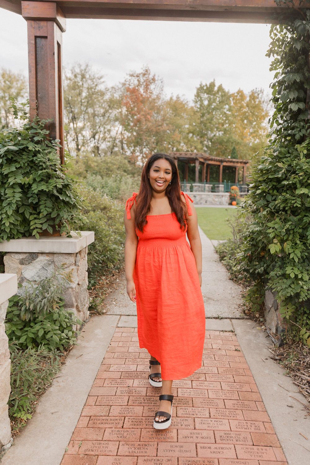 Woman in bright coral summer dress walks along brick pathway surrounded by greenery in outdoor garden setting.