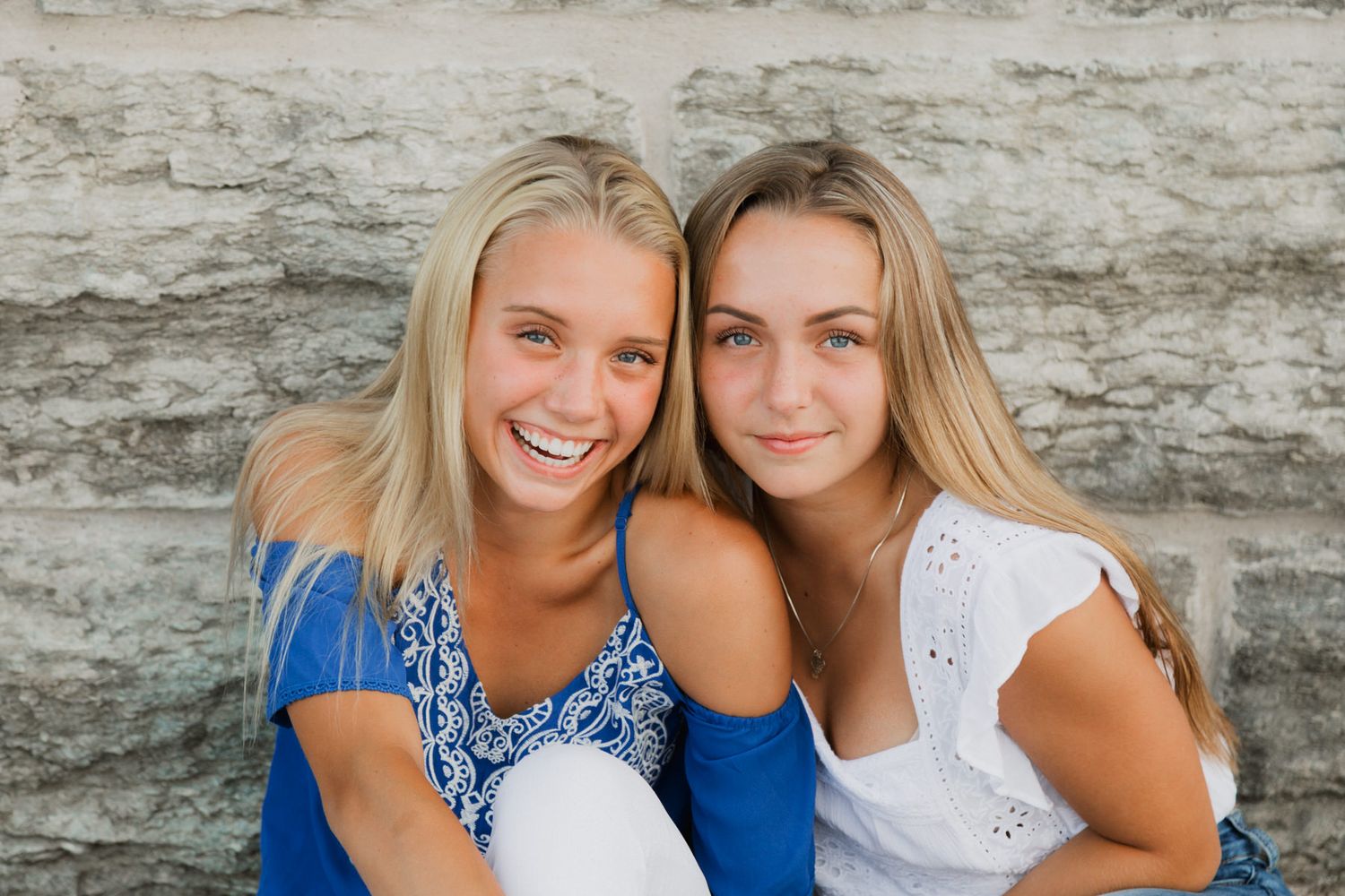 Two smiling blonde teenage girls in blue and white tops posing together against a stone wall backdrop.