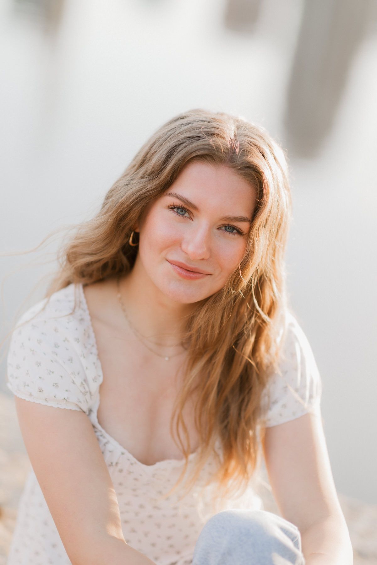 Young blonde woman with blue eyes in floral top smiling softly in bright natural light.