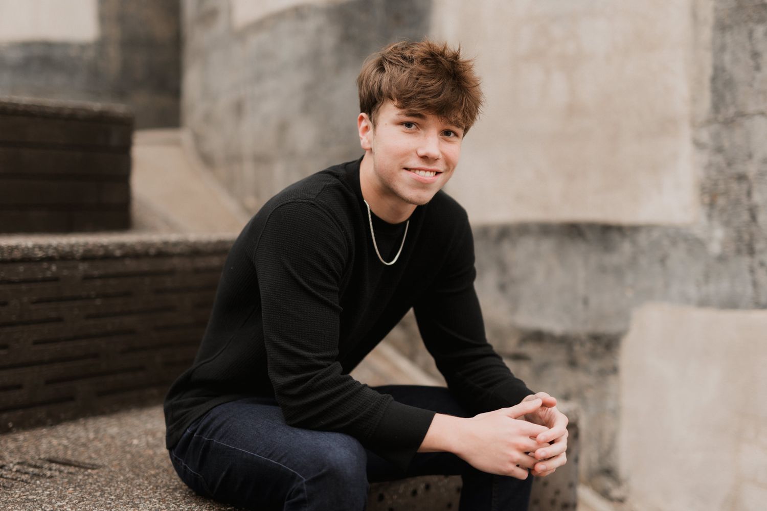 Young man in black sweater sitting on stone steps outdoors with a silver chain necklace.