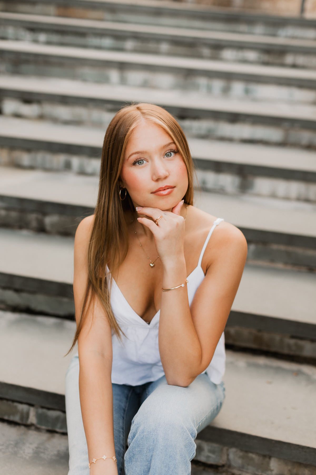 Young woman with long hair sits on stone steps, wearing white camisole and jeans, resting chin on hand.
