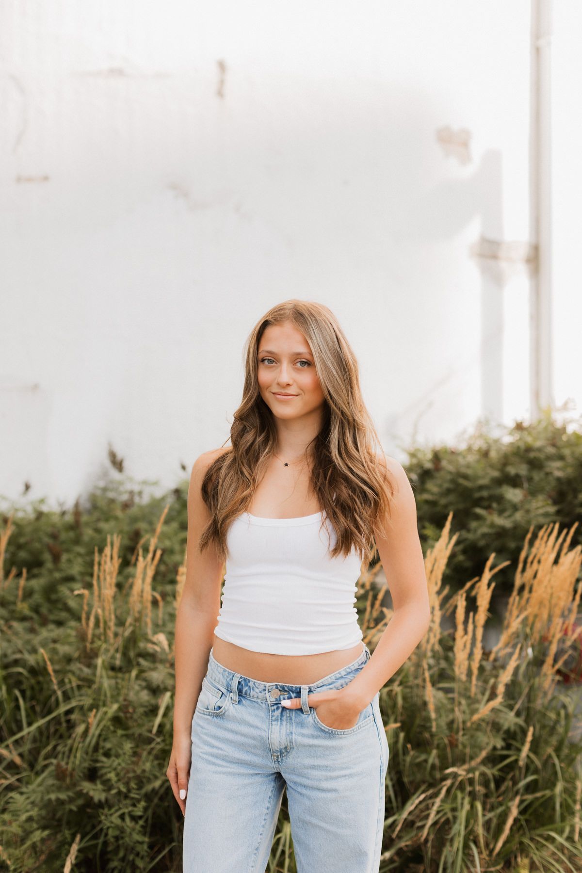 Teen girl in white crop top and jeans stands confidently in front of lush greenery.
