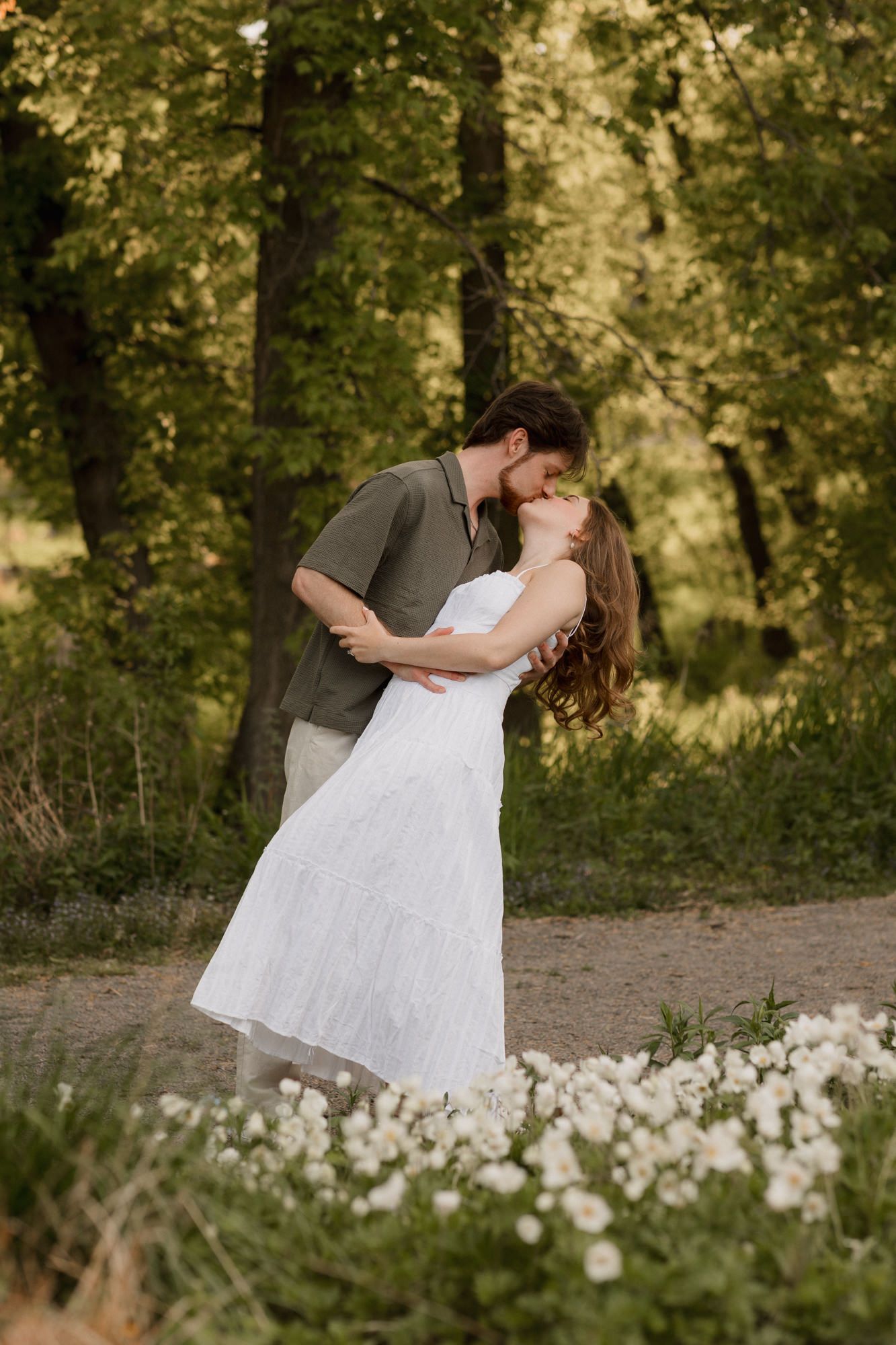 Couple sharing a romantic kiss in a lush green forest setting, woman in white dress surrounded by wildflowers.