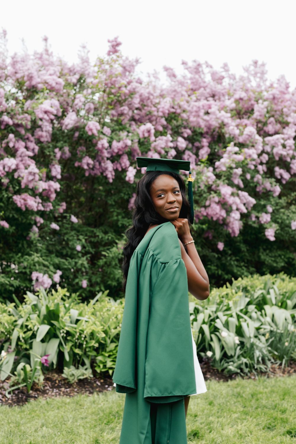 Graduate in emerald academic regalia stands beside flowering spring bushes.