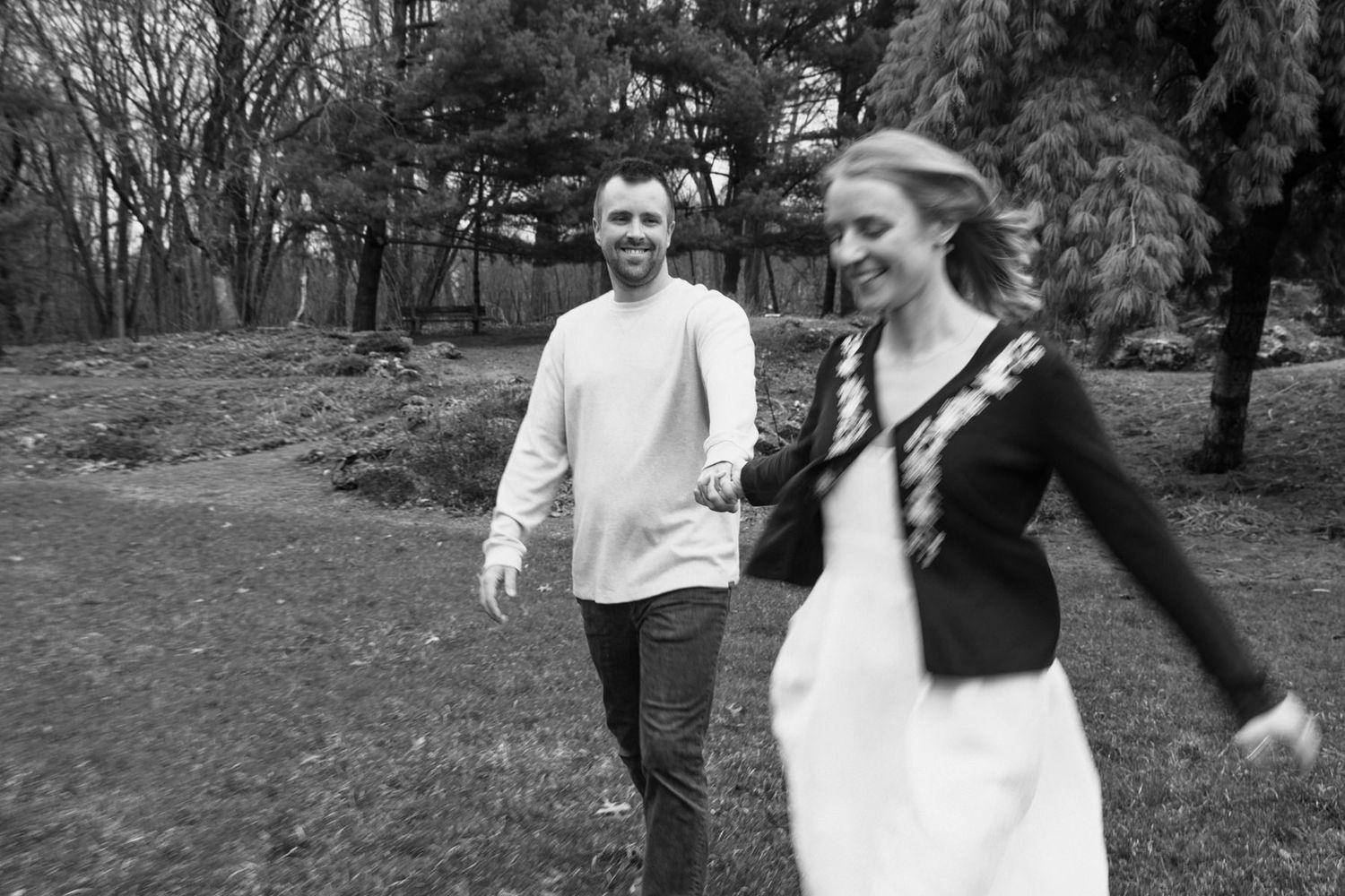 Black and white photo of a smiling couple holding hands and walking through a lush green park together.