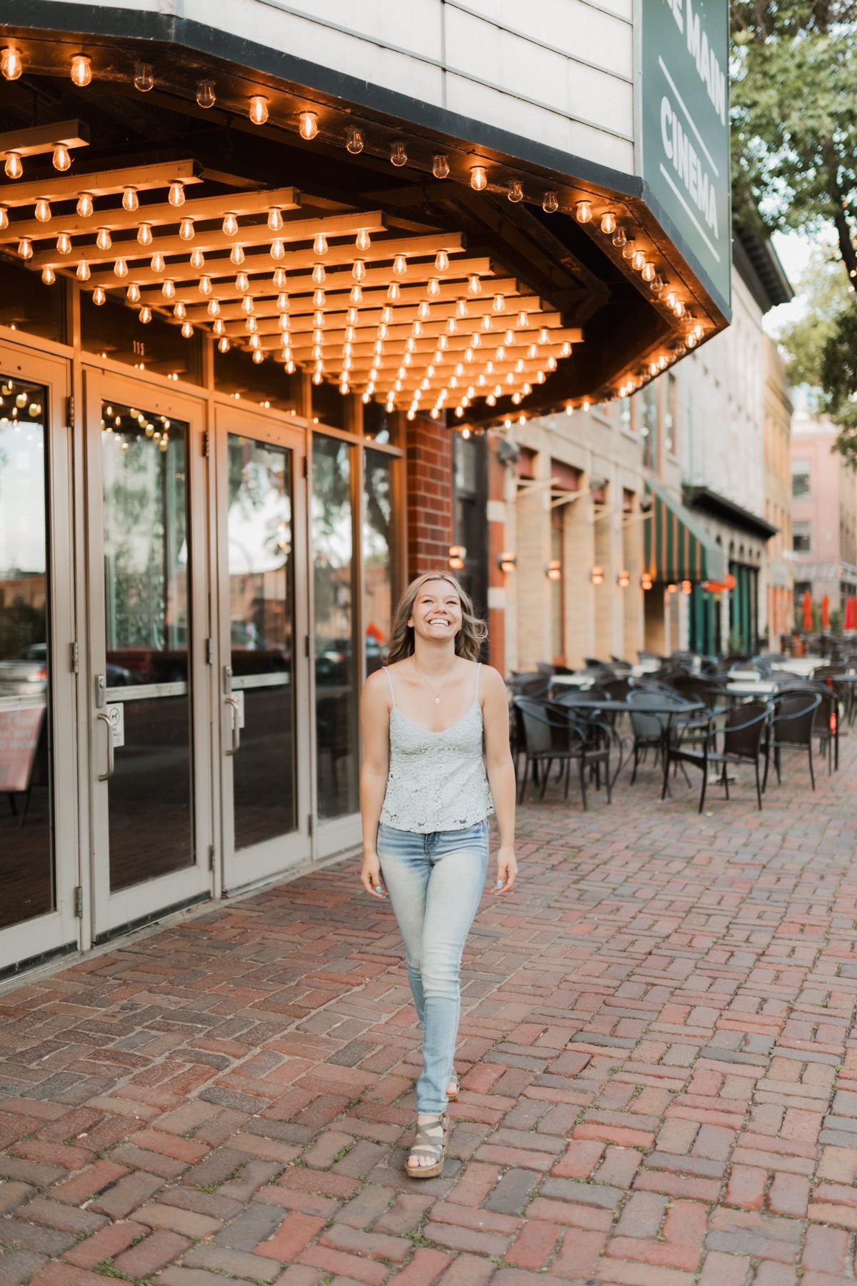 Young woman in jeans and gray tank top smiling while walking under lit marquee lights outside a brick building.