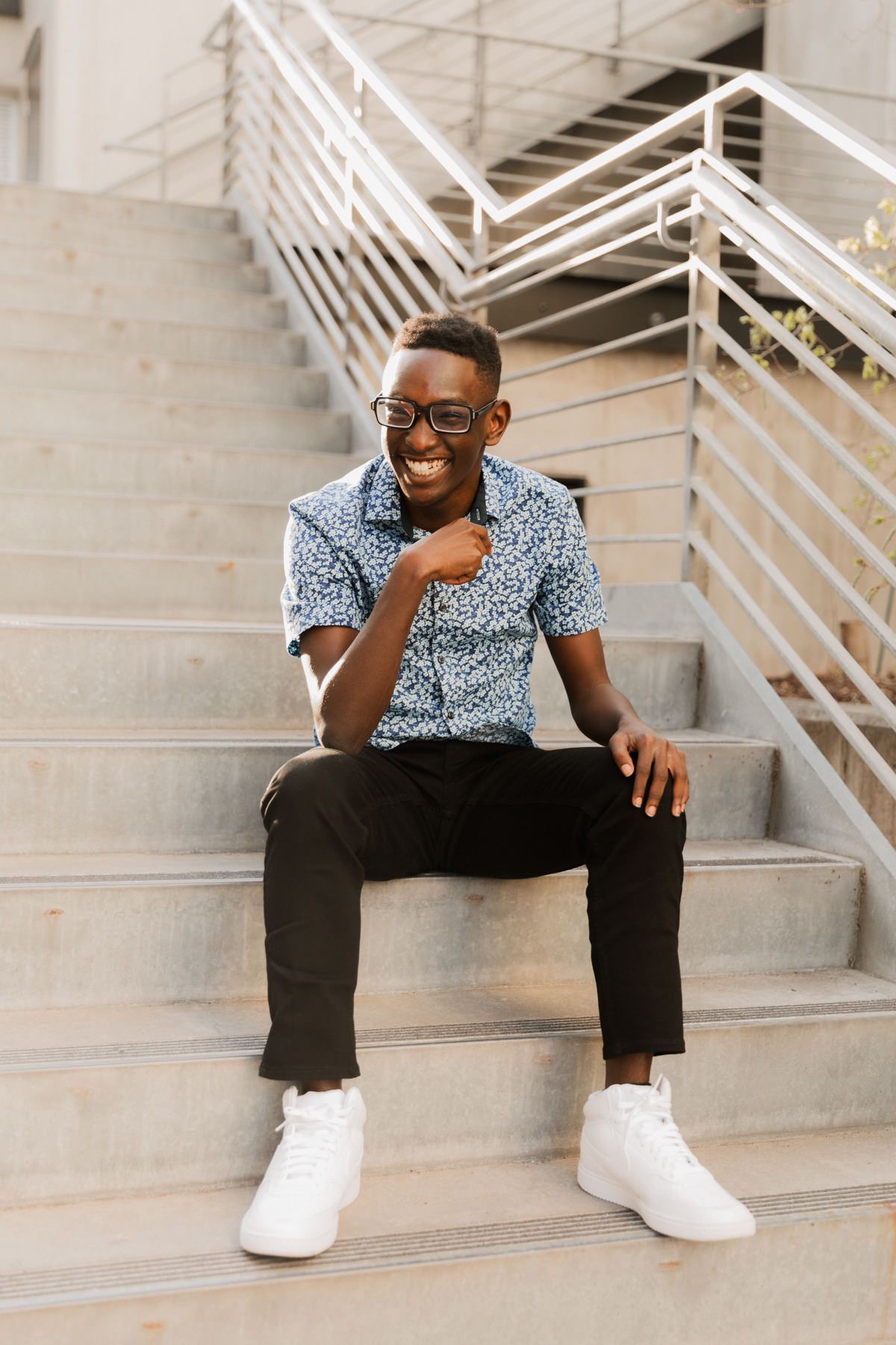 Smiling young Black man in floral shirt and glasses sitting on outdoor concrete steps.