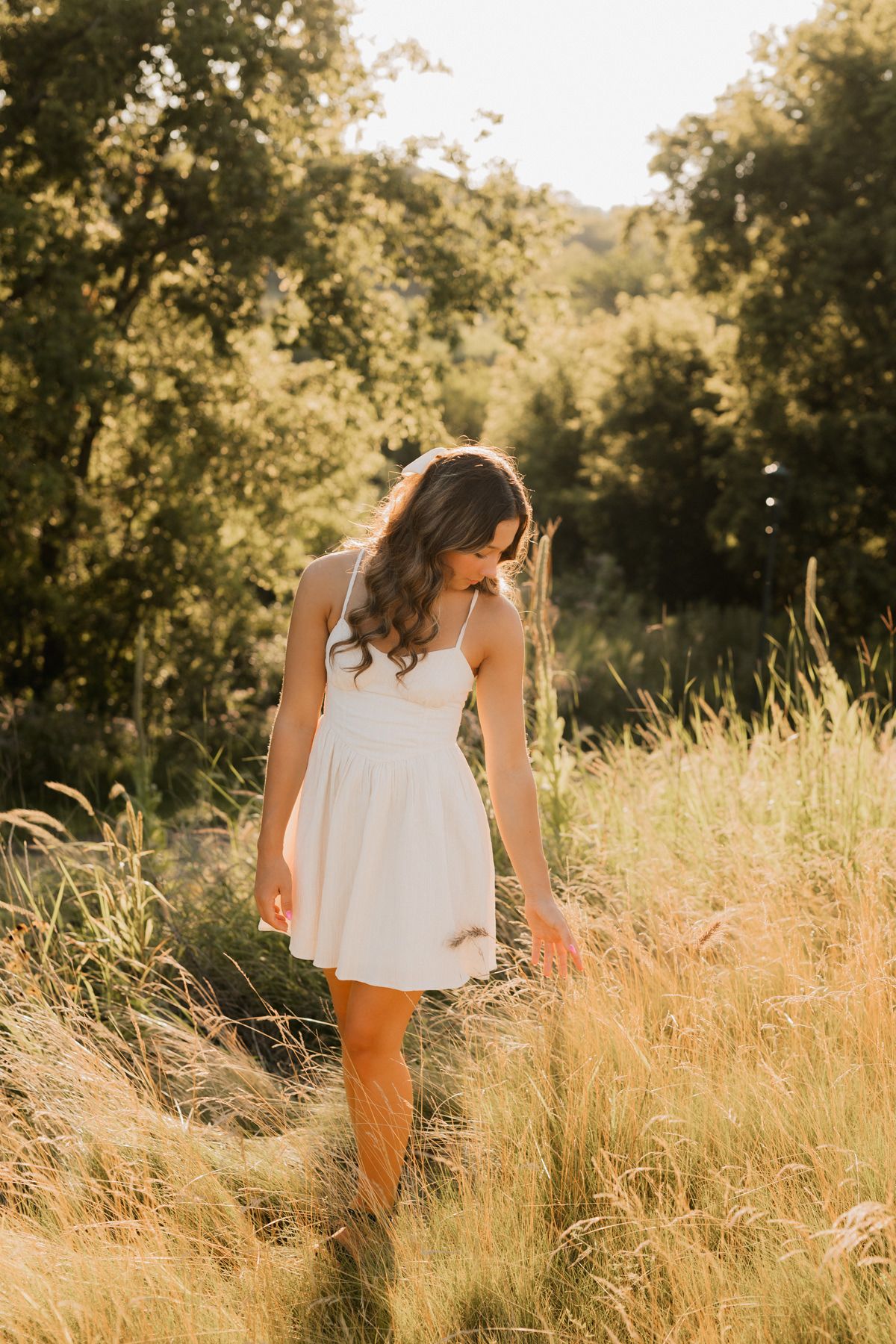 Young woman in white sundress walks through golden tall grass field with lush green trees in warm sunlight.
