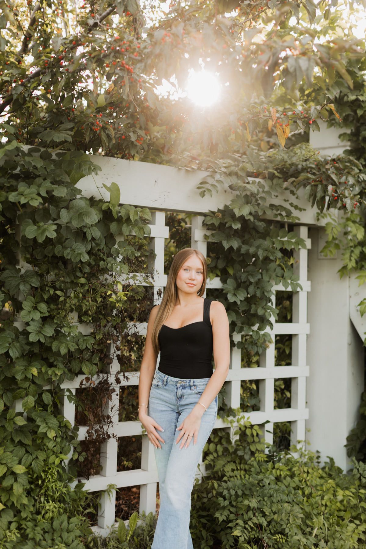 Blonde woman in black tank top and jeans stands before a white garden trellis covered in green vines.