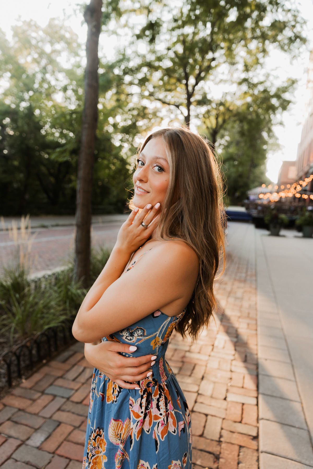 Smiling brunette woman in a floral strapless dress poses on a brick sidewalk at golden hour.