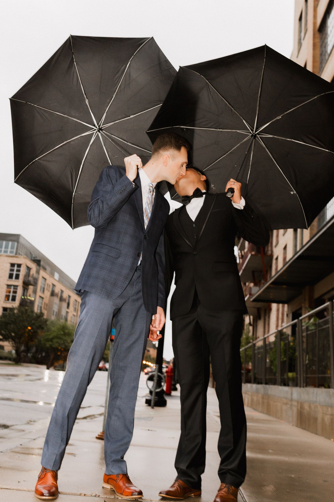 Two grooms in suits share a kiss under black umbrellas on a rainy wedding day outdoors.