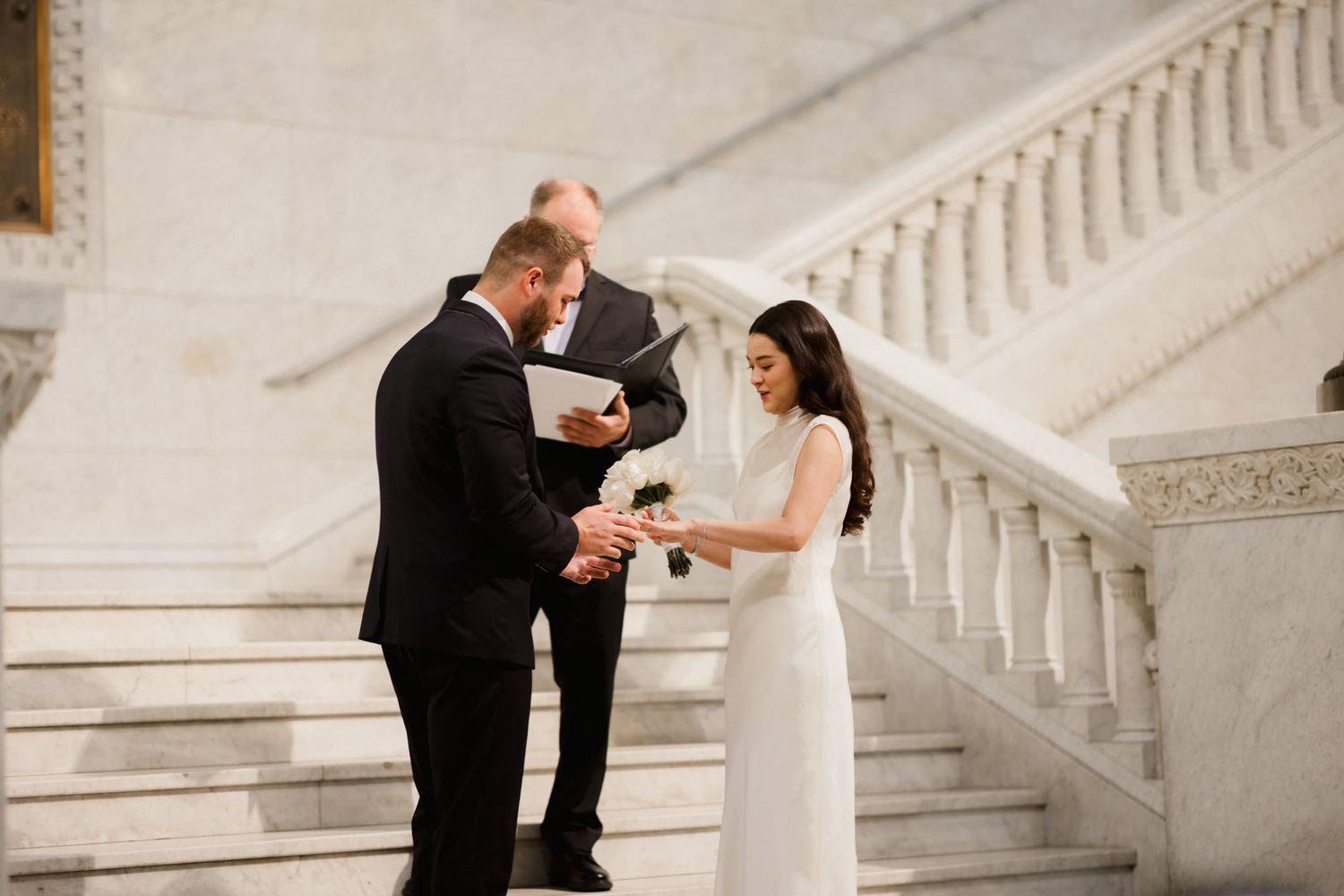 Couple exchanging vows at elegant indoor staircase wedding ceremony with officiant holding book.