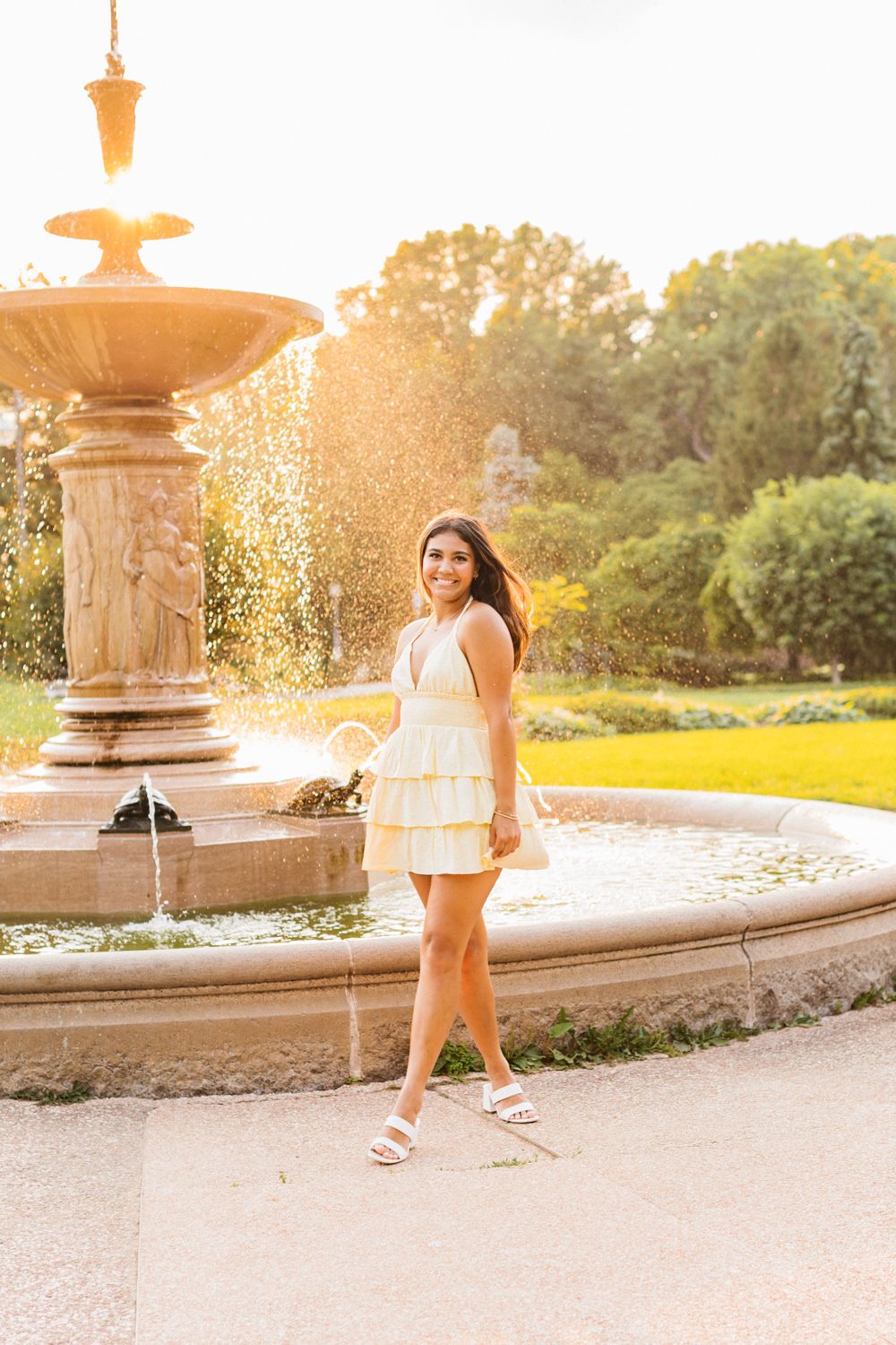 Someone in a white dress poses gracefully by an ornate fountain at sunset creating a dreamy summer atmosphere.