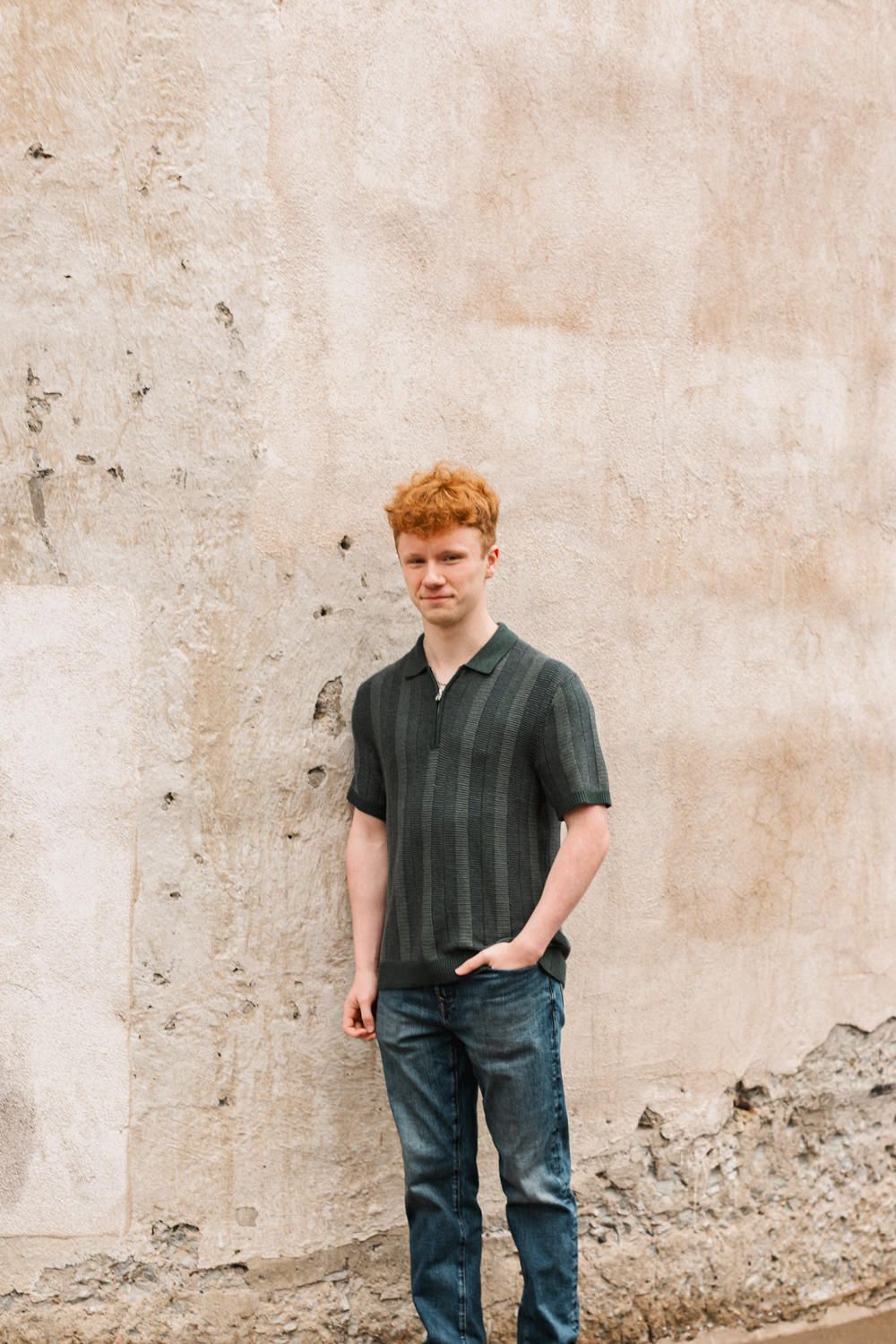 Person standing against a beige textured wall wearing dark shirt and jeans in an outdoor setting.