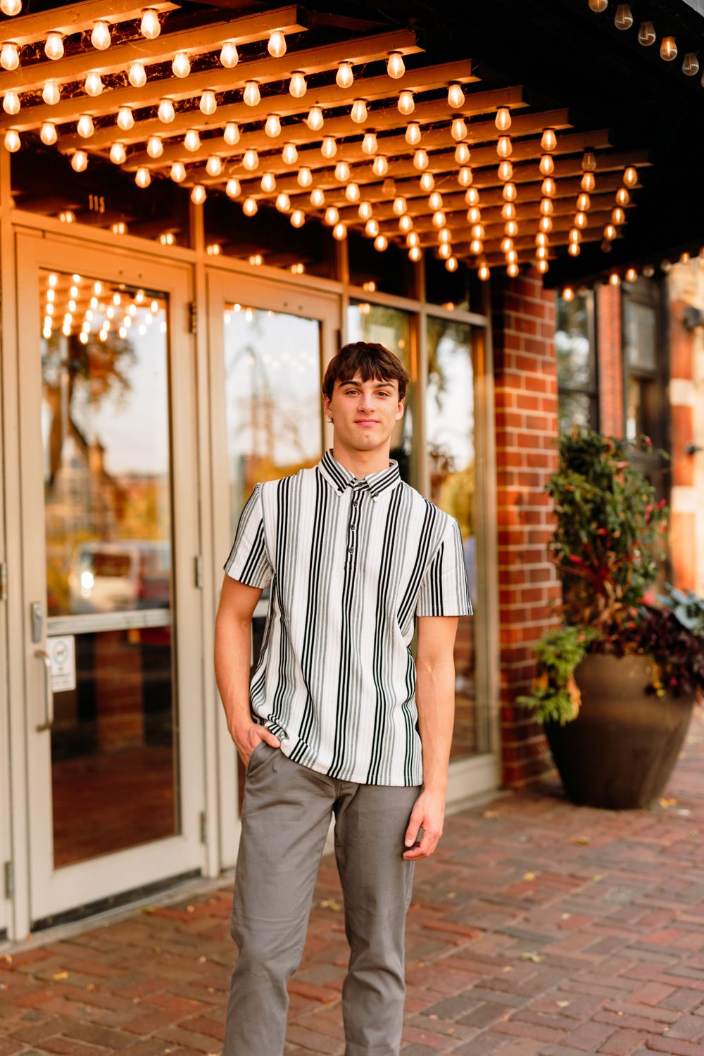 Person in striped shirt poses by brick wall and string lights in outdoor setting.