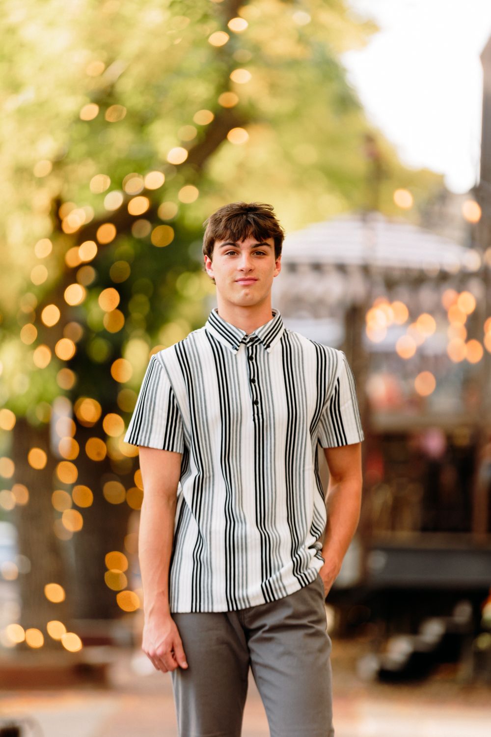 A person in a striped shirt poses outdoors with bokeh lights and trees creating a warm evening atmosphere.