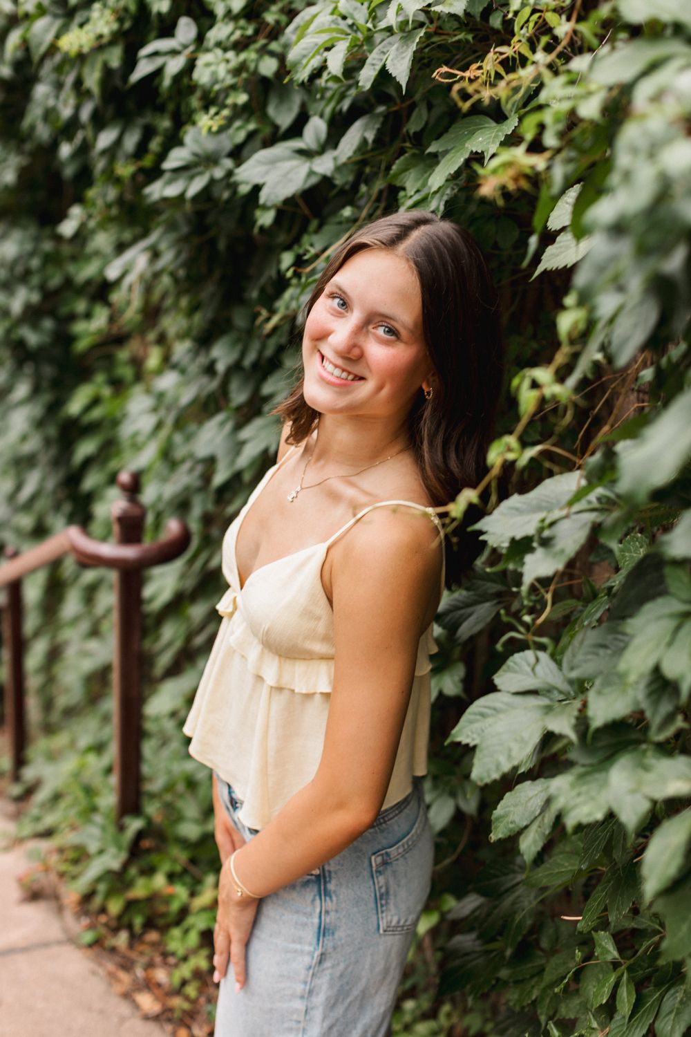 A joyful portrait against a backdrop of lush green vines, wearing a cream-colored top and light denim jeans.
