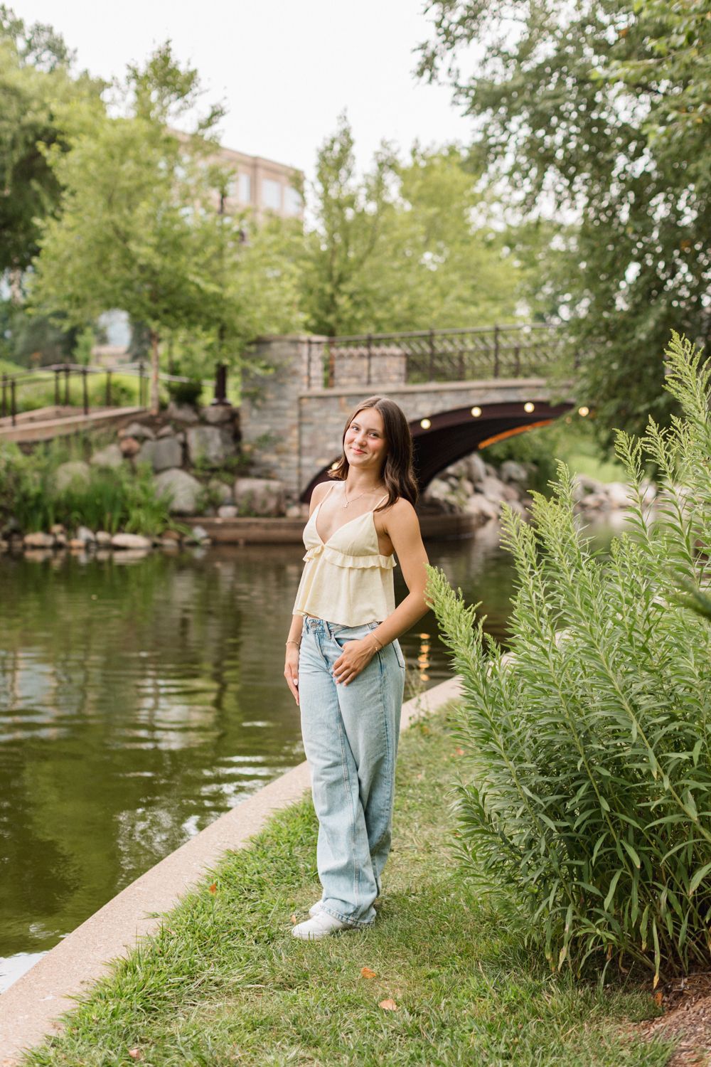 A person in a beige top and light jeans stands along a tranquil pond with a stone bridge and lush greenery.