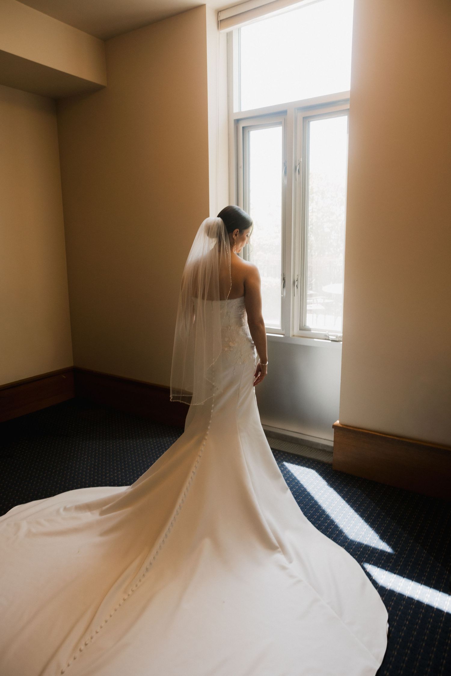 Bride in white wedding gown with long veil and train stands by sunlit window in elegant room.