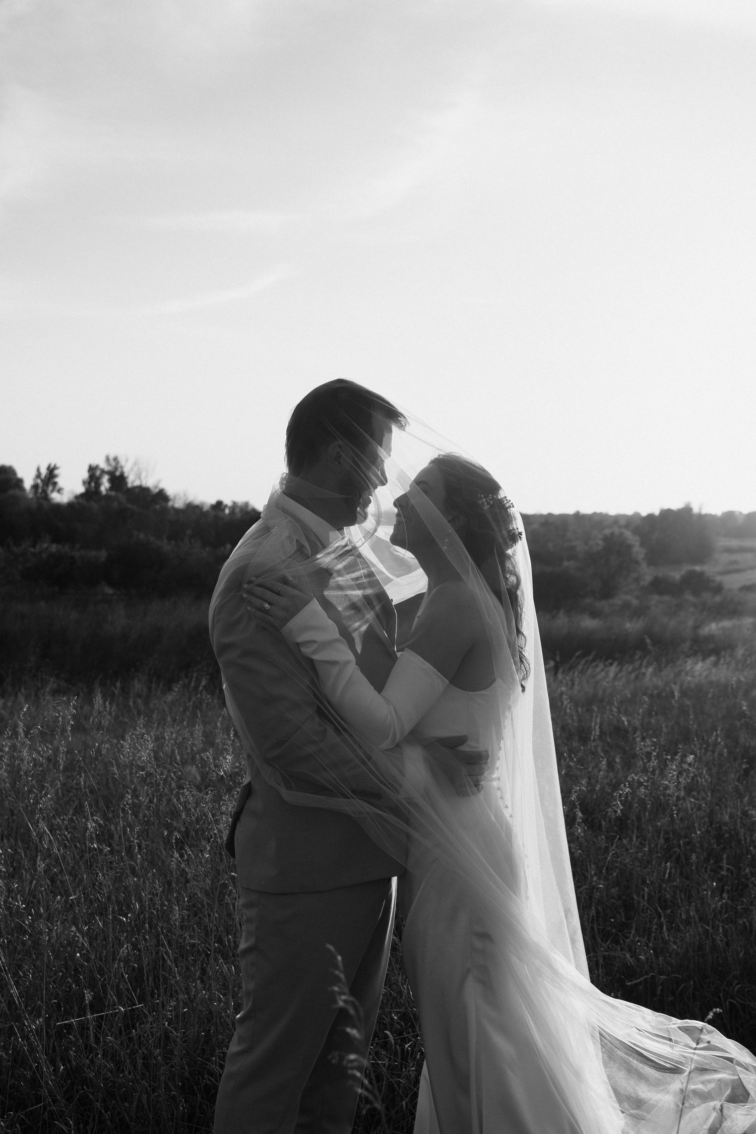 Black and white photo of a bride and groom embracing face to face under a sheer veil in a grassy field at sunset.