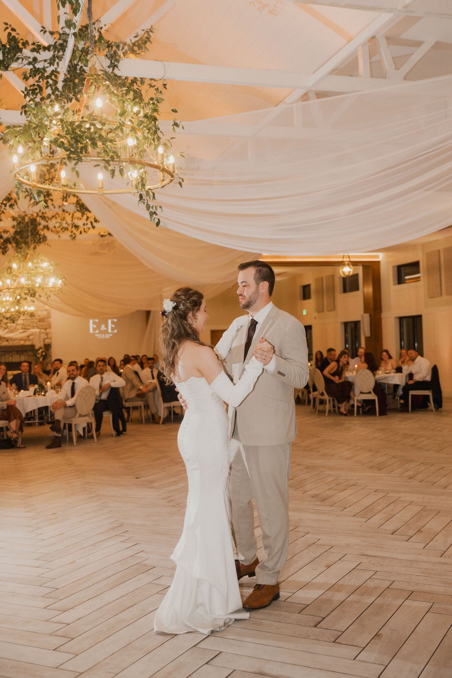 Bride and groom share a first dance on a wooden floor beneath draped fabric and greenery chandeliers as guests watch.
