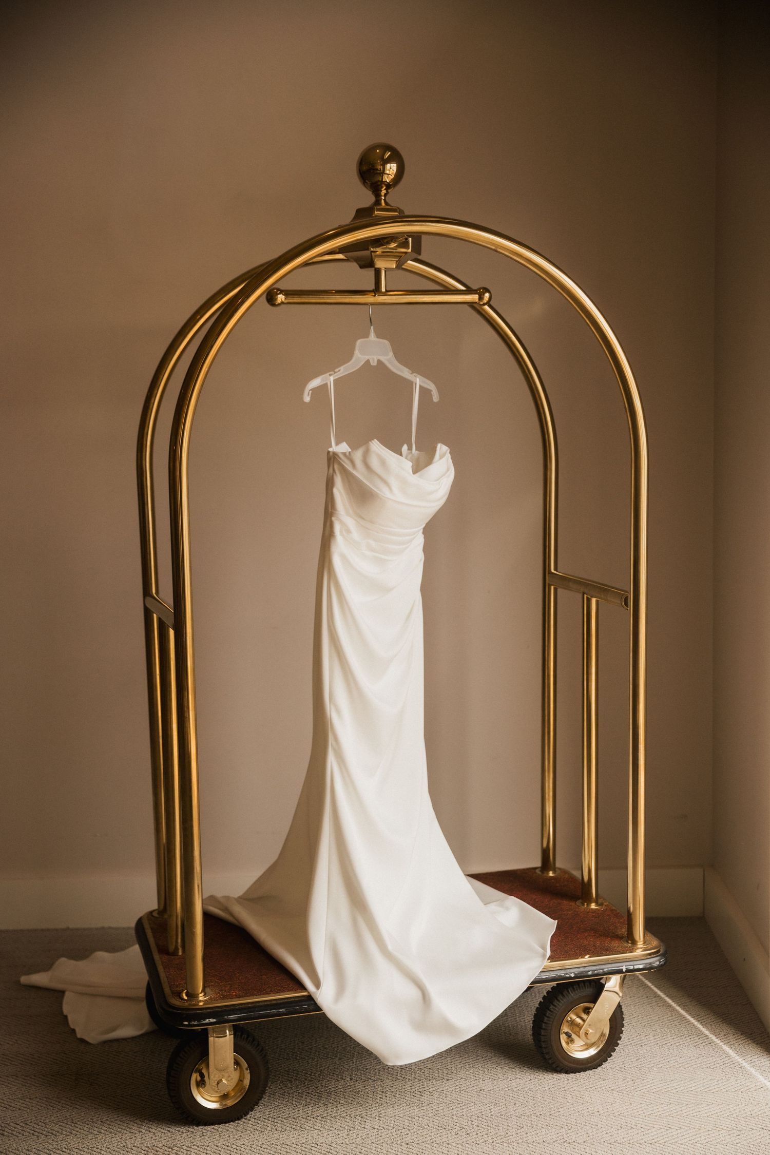 White strapless wedding gown hanging on a vintage brass luggage cart in an elegant hotel room.