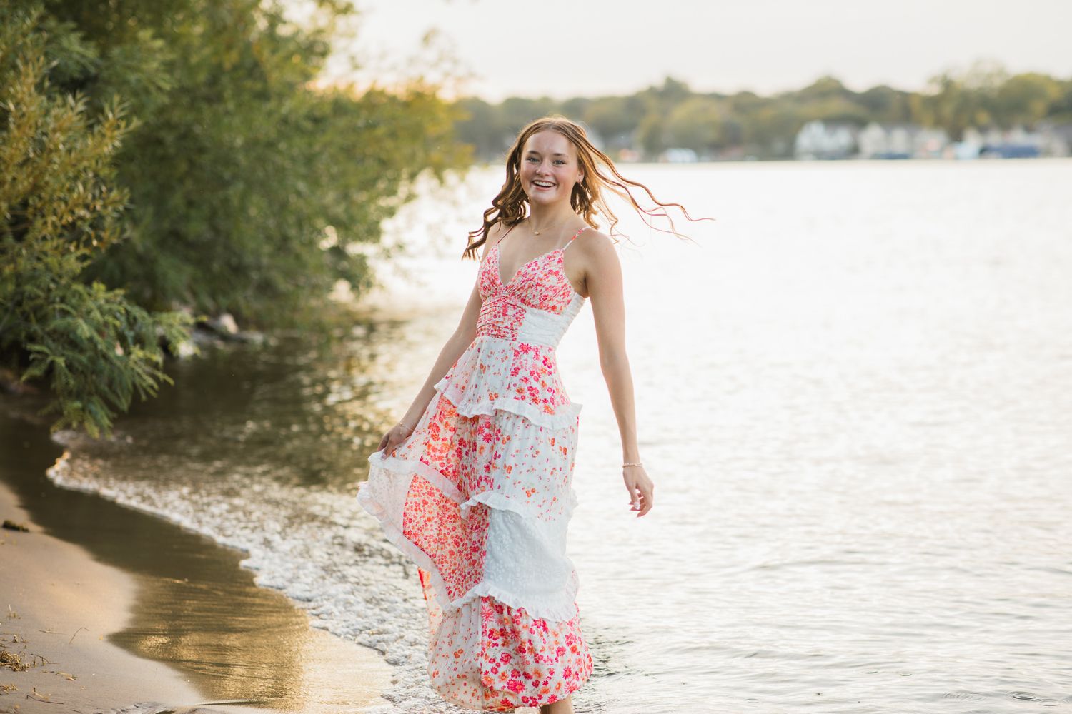 A joyful young woman in a floral tiered maxi dress walks along a sunlit lakeshore with trees in the background.