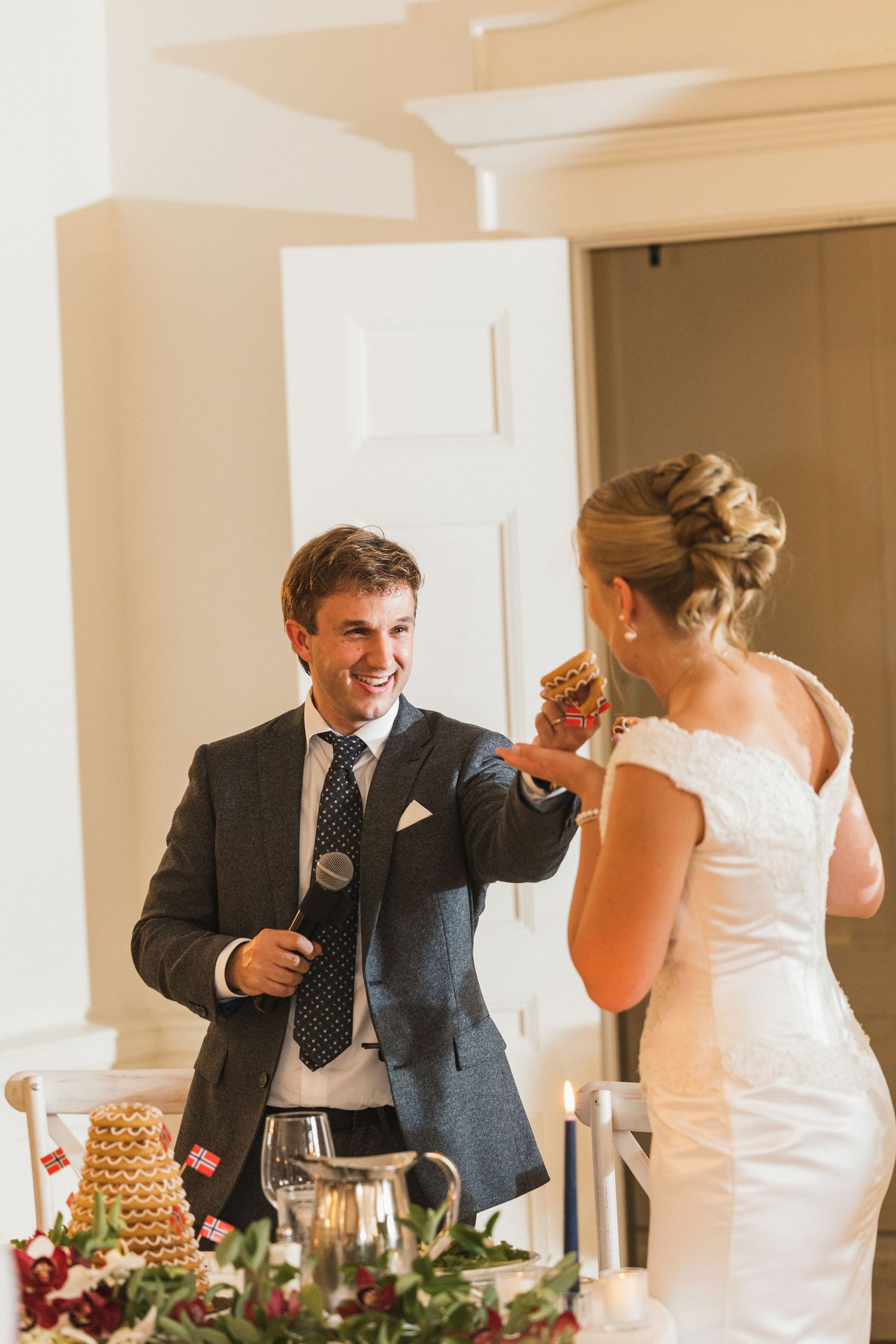Smiling groom holds a piece of kransekake cake toward the bride at their elegantly decorated wedding reception table.