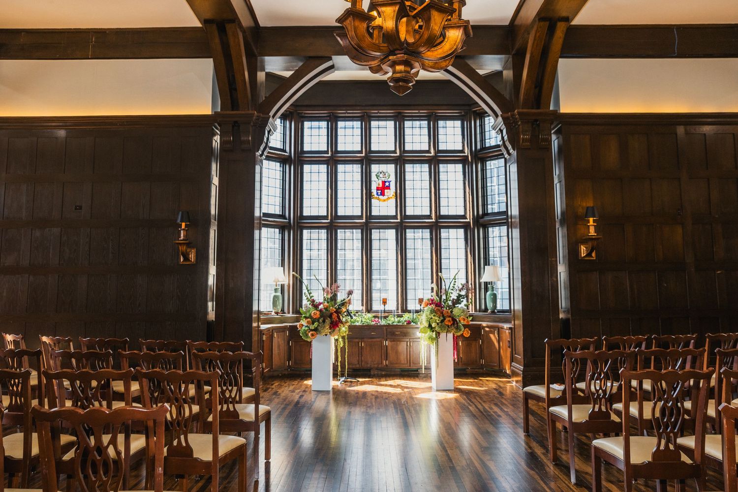 Elegant wedding ceremony setup in a dark wood-paneled room with floral arrangements flanking a grand arched window.