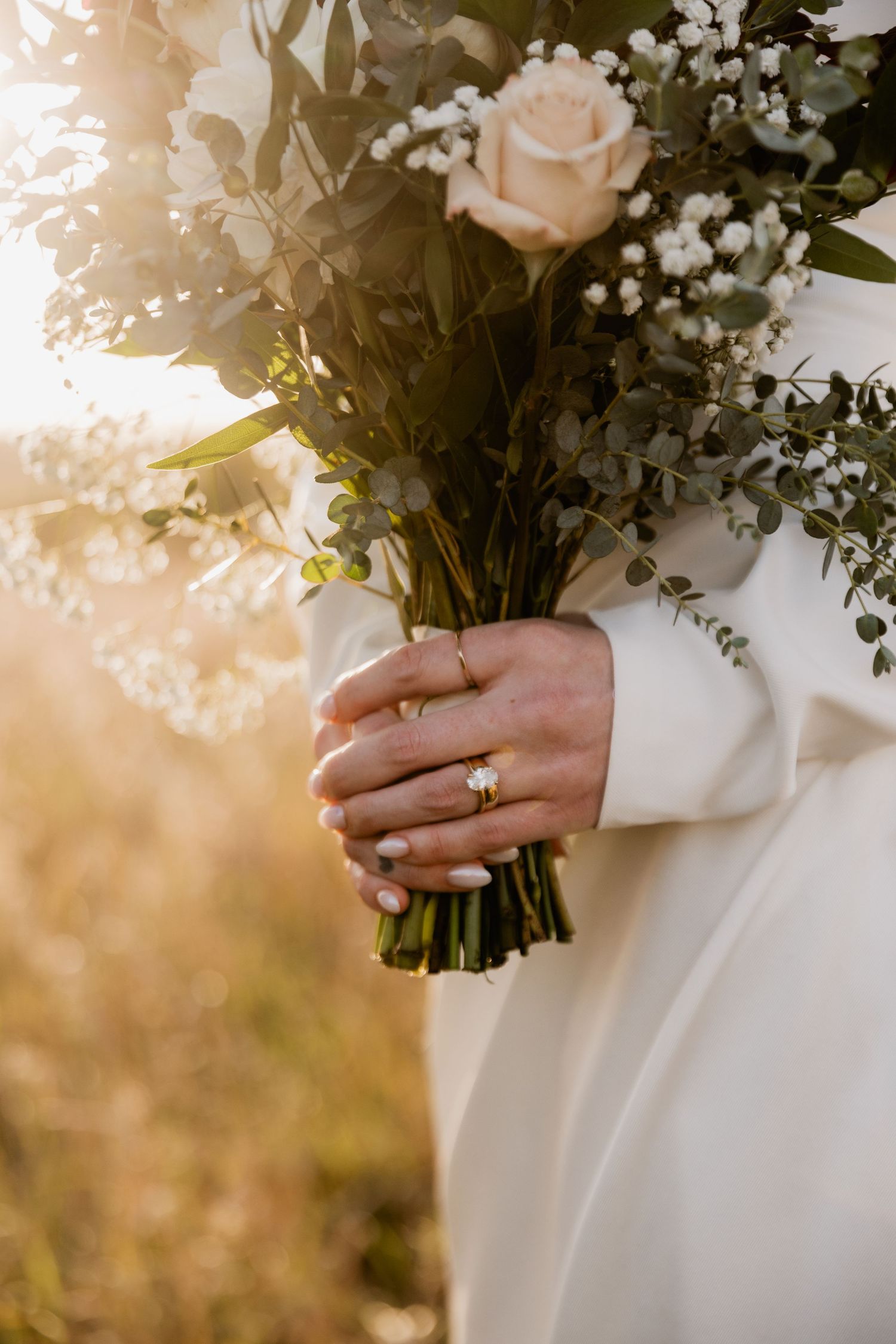 Bride holding a wedding bouquet with roses and greenery, wearing a gold engagement ring, in golden sunlight.