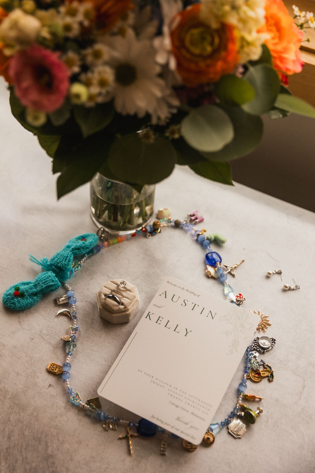 Wedding details flat lay with charm bracelet, rings, and Austin & Kelly program book beside a colorful floral arrangement.