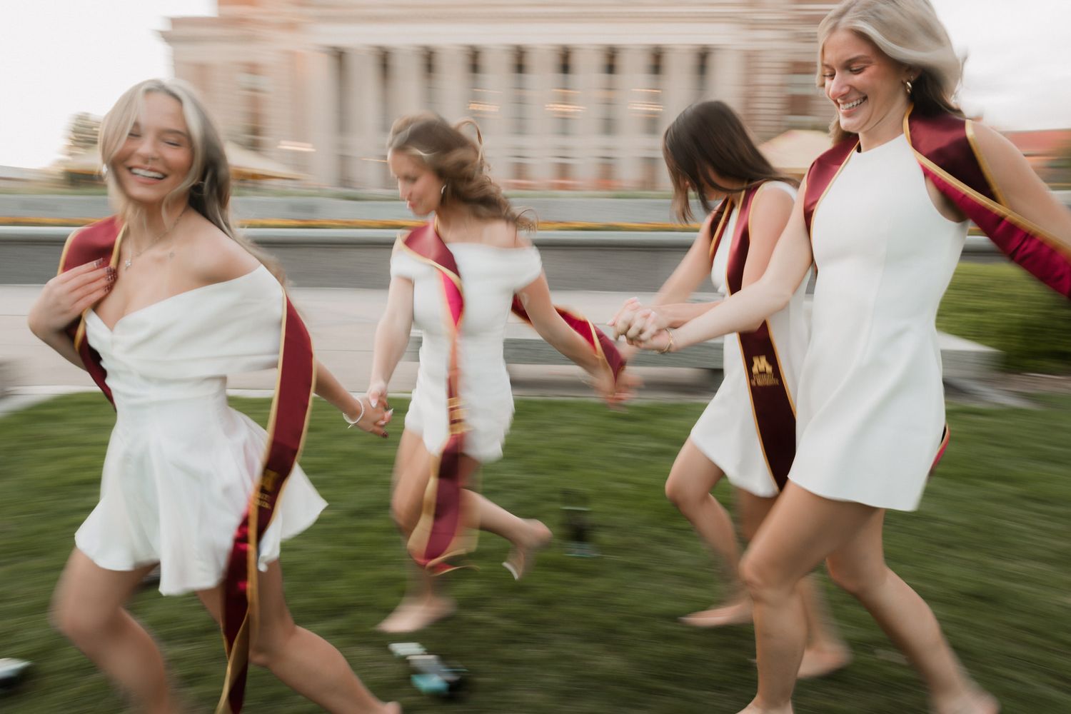 Four women in white dresses and maroon graduation stoles run holding hands on a green lawn.