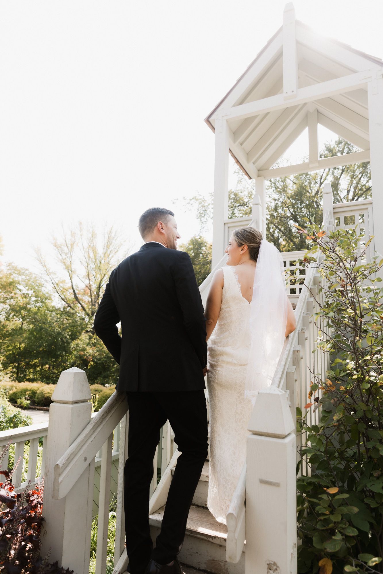 A bride in a white gown and veil stands with a groom in a black suit on a white gazebo staircase surrounded by lush greenery.
