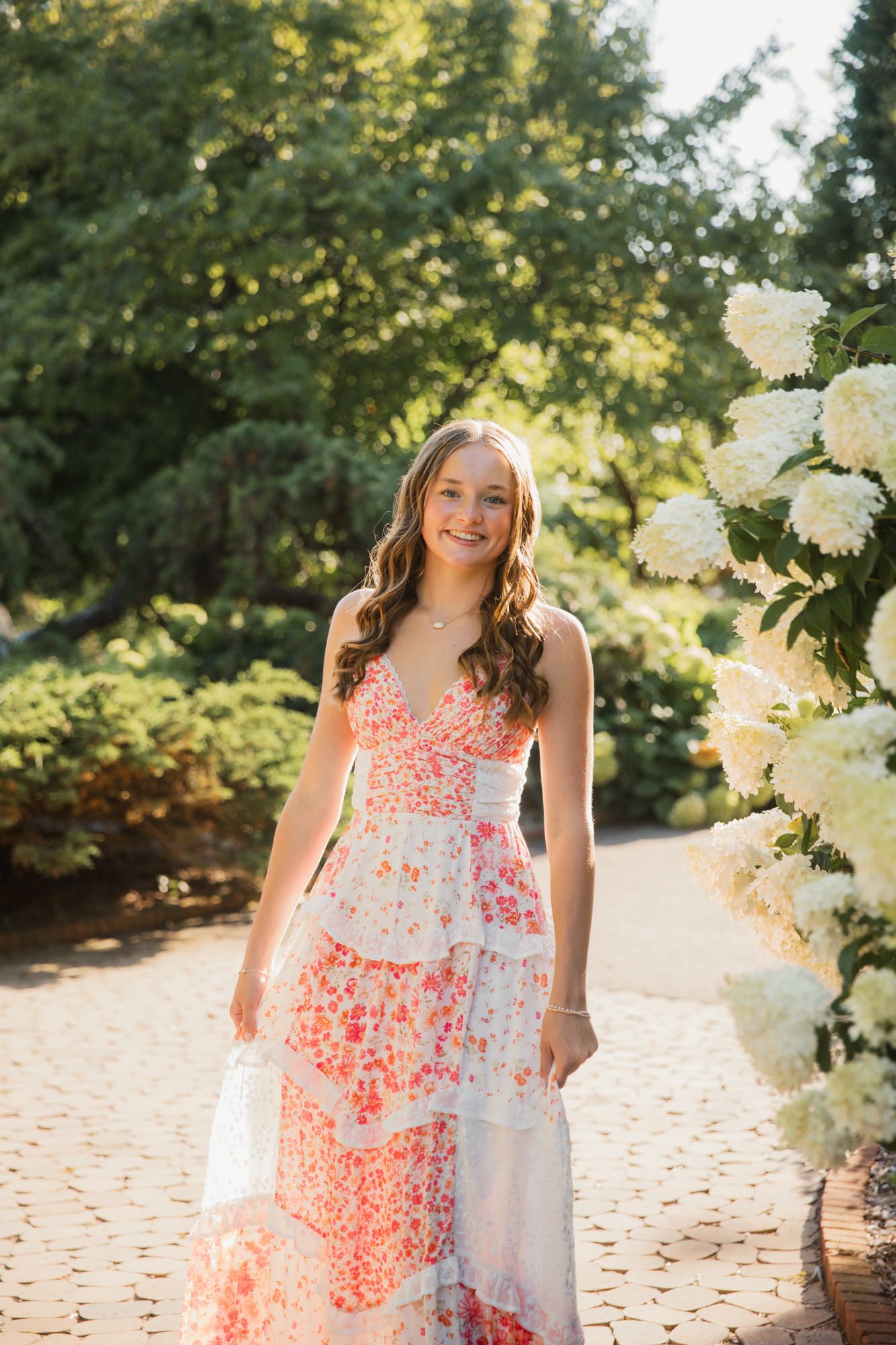 Young woman smiling in a floral tiered maxi dress, standing in a sunny garden with white hydrangeas.