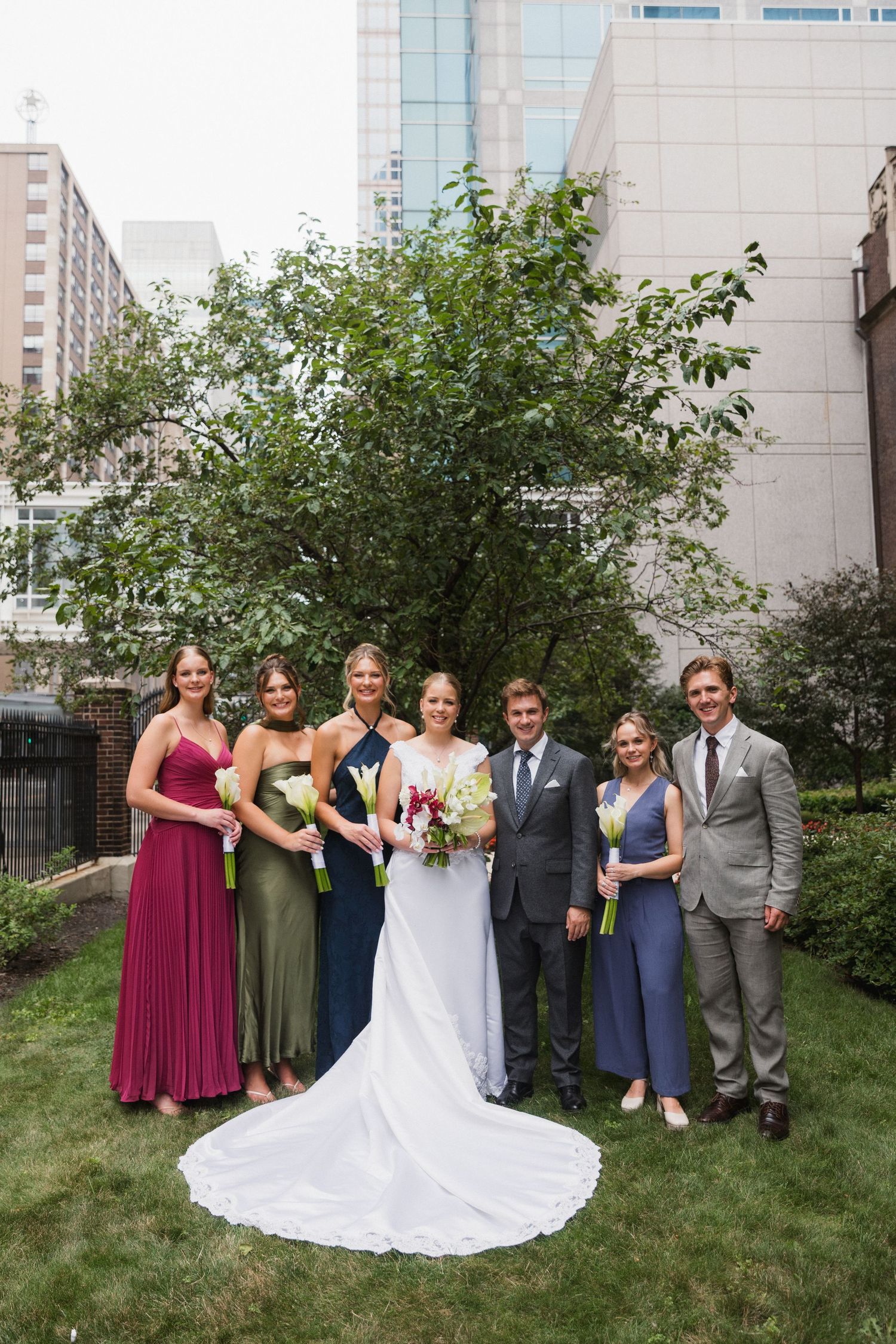 Wedding party poses outdoors in urban garden, bride in white gown with bridesmaids in colorful dresses and groomsmen in suits.