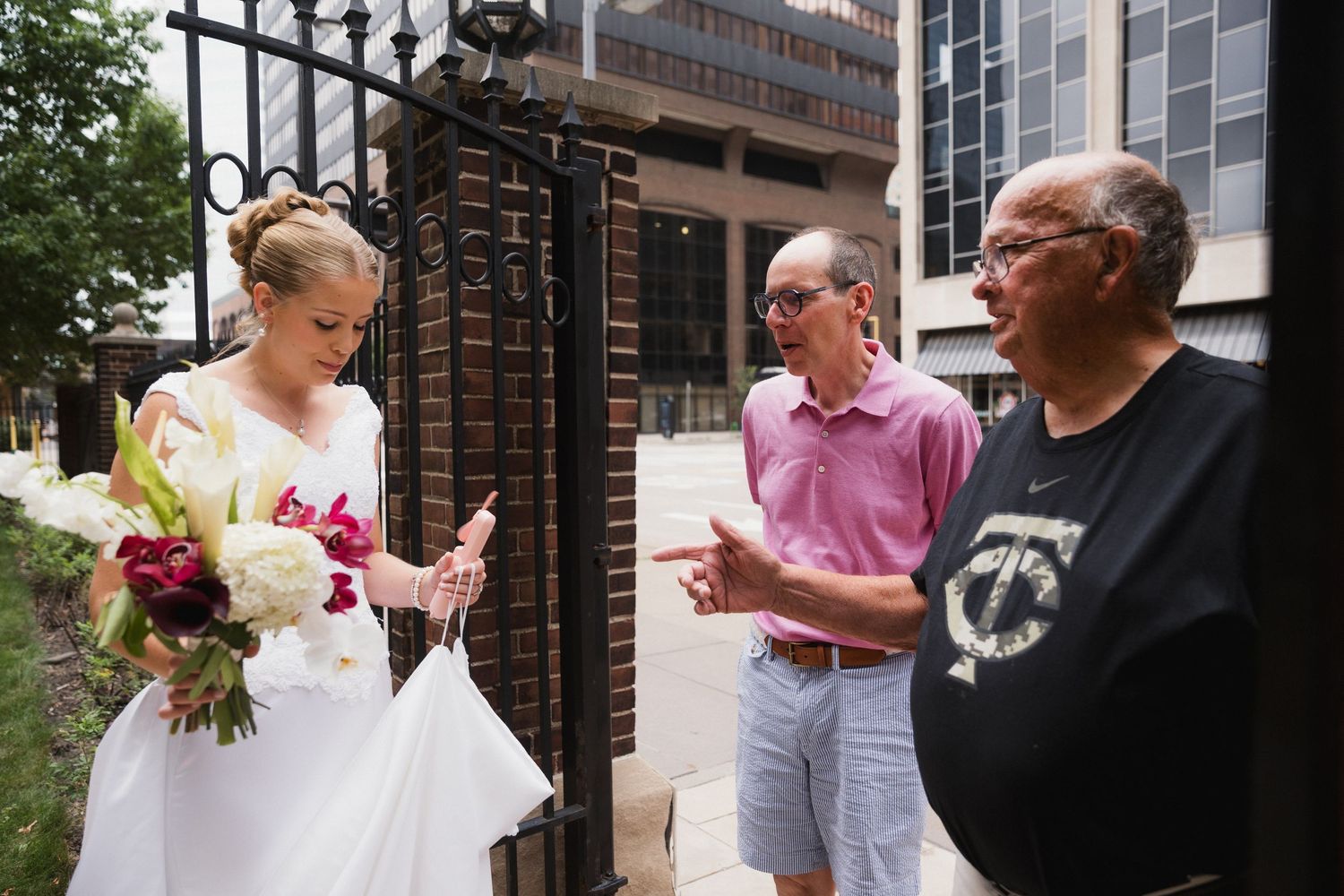 Bride in white gown holds bouquet while conversing with two men near ornate iron gates outside a building.