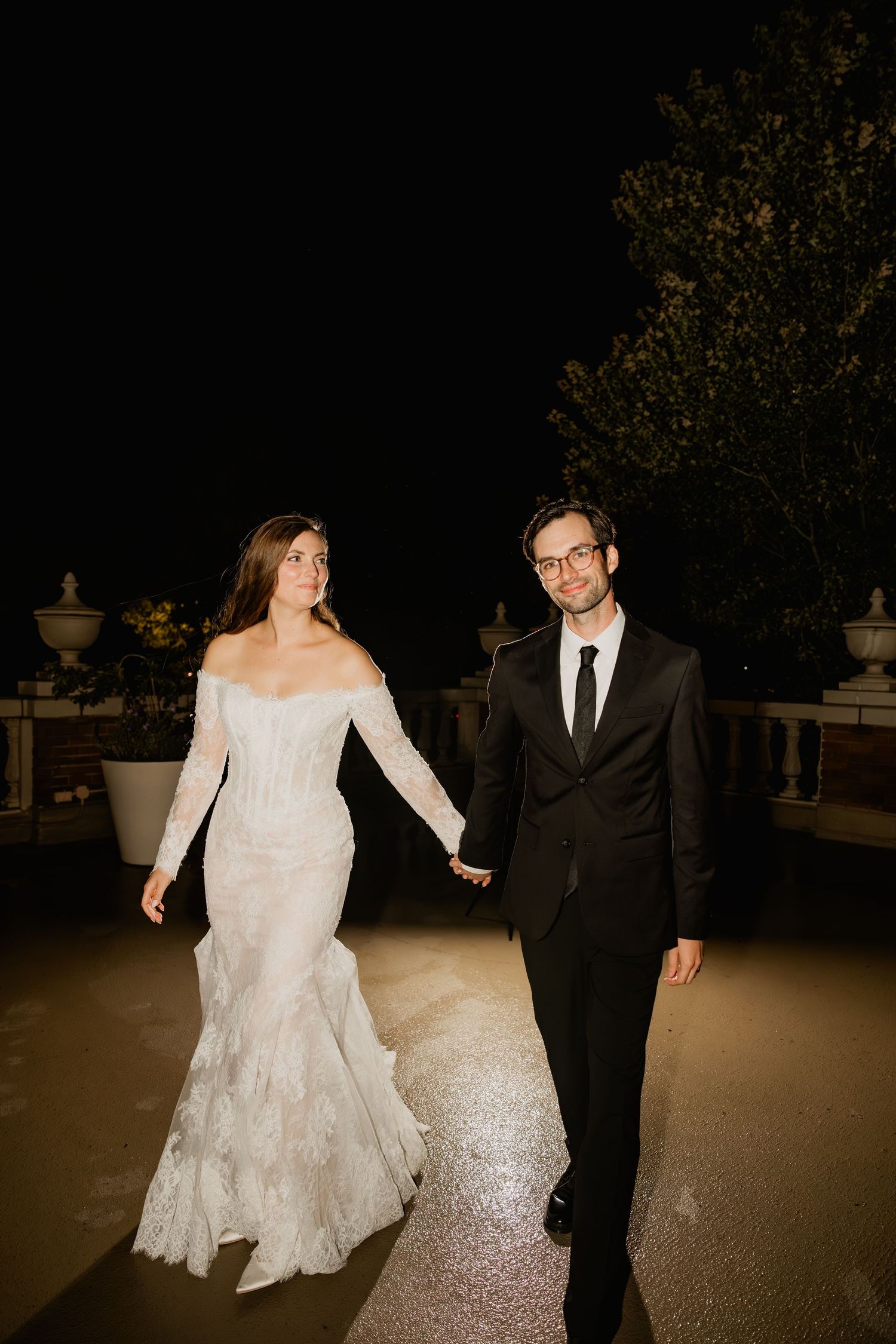 Bride in off-shoulder lace gown and groom in black suit hold hands walking outdoors at night.