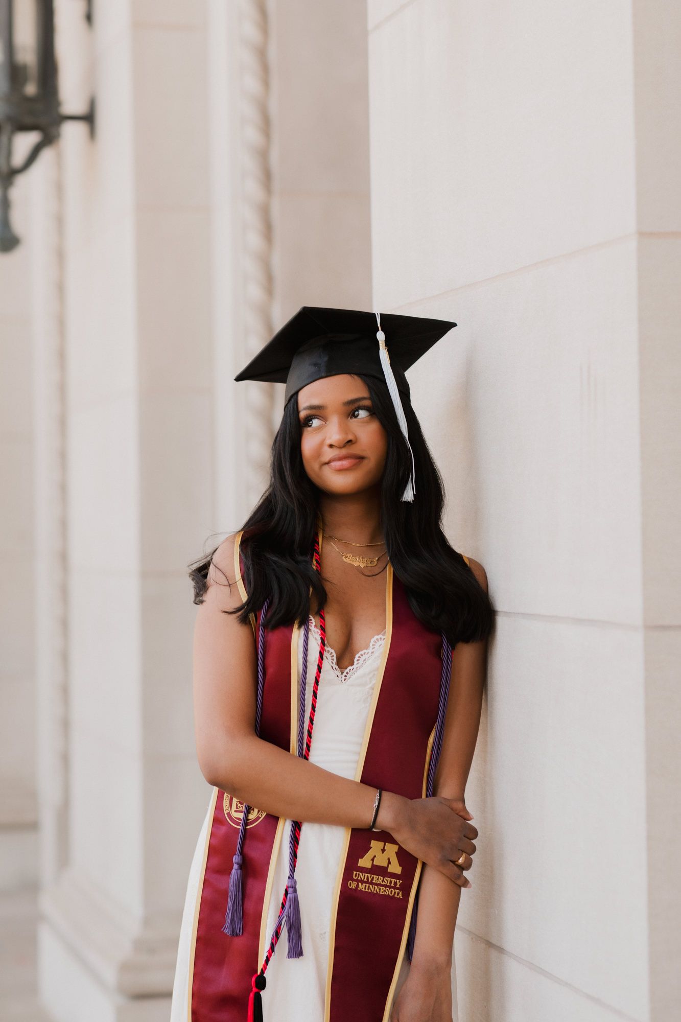 Smiling graduate in black cap and maroon University of Minnesota stole poses against stone column outdoors.