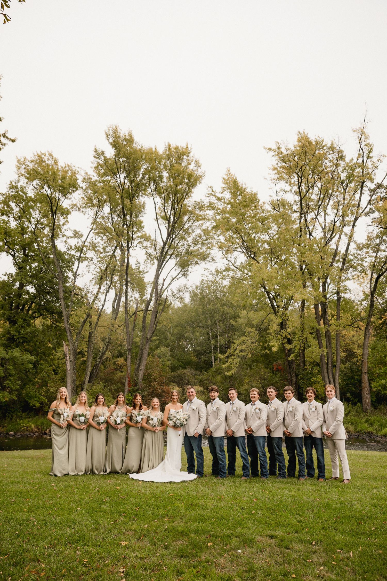 Wedding party in neutral tones standing on green lawn with trees, bridesmaids in sage dresses and groomsmen in beige suits.